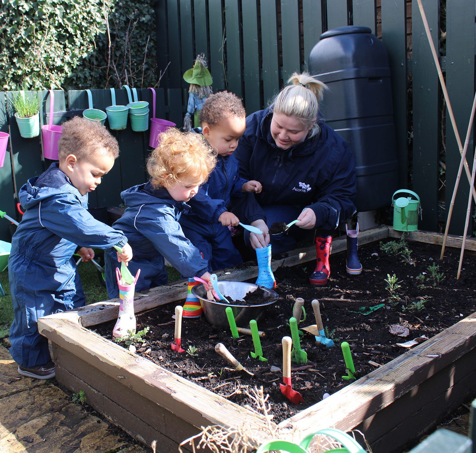 Acorns Nurseries Adults and children in blue coats planting seeds in a garden bed, with gardening tools and colorful watering cans nearby, nurturing their efforts like tender acorns ready to grow.