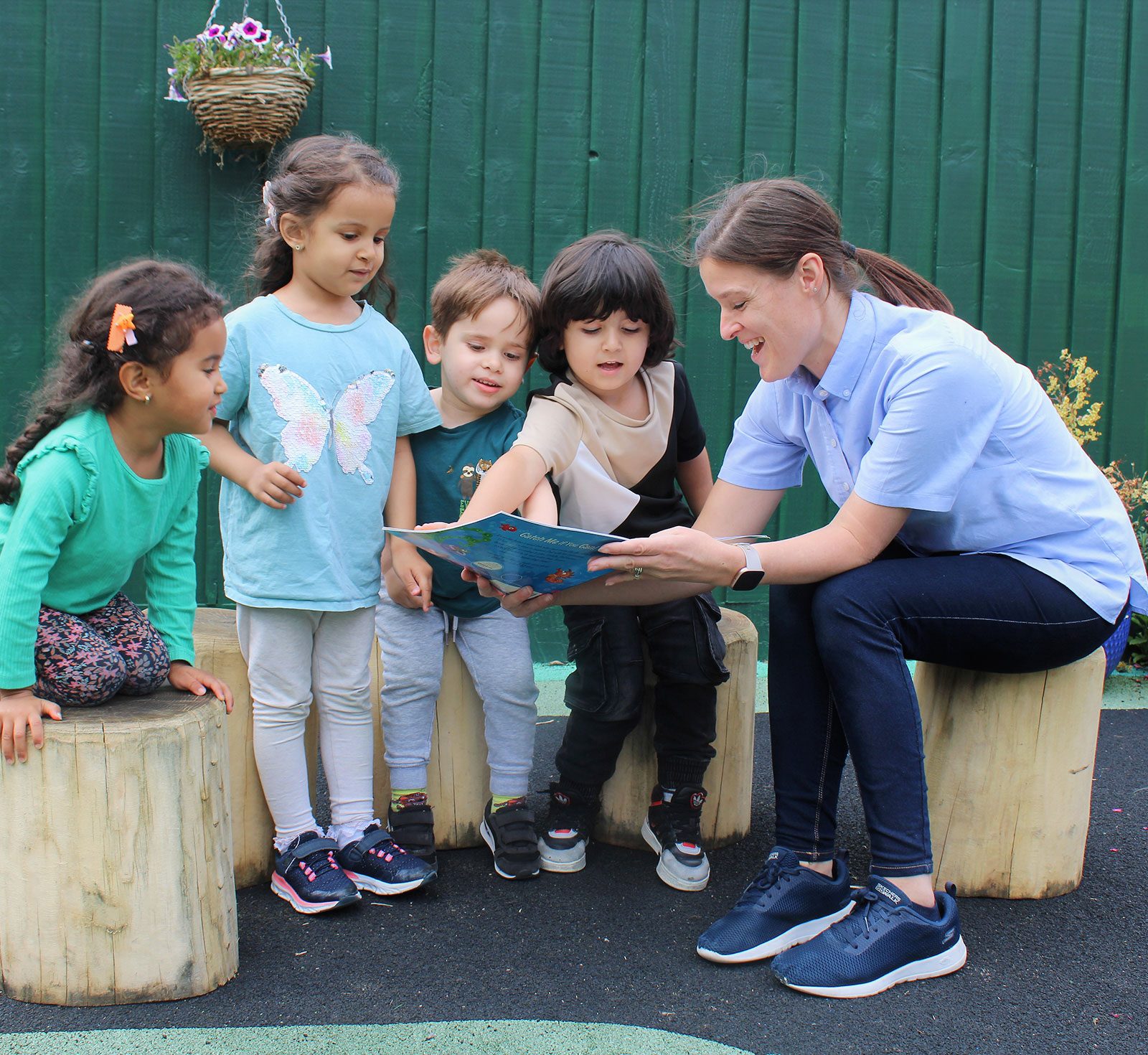 Acorns Nurseries A woman reads a book to four children sitting on wooden stools outside Acorns House, their imaginations taking flight against the backdrop of a vibrant green fence.