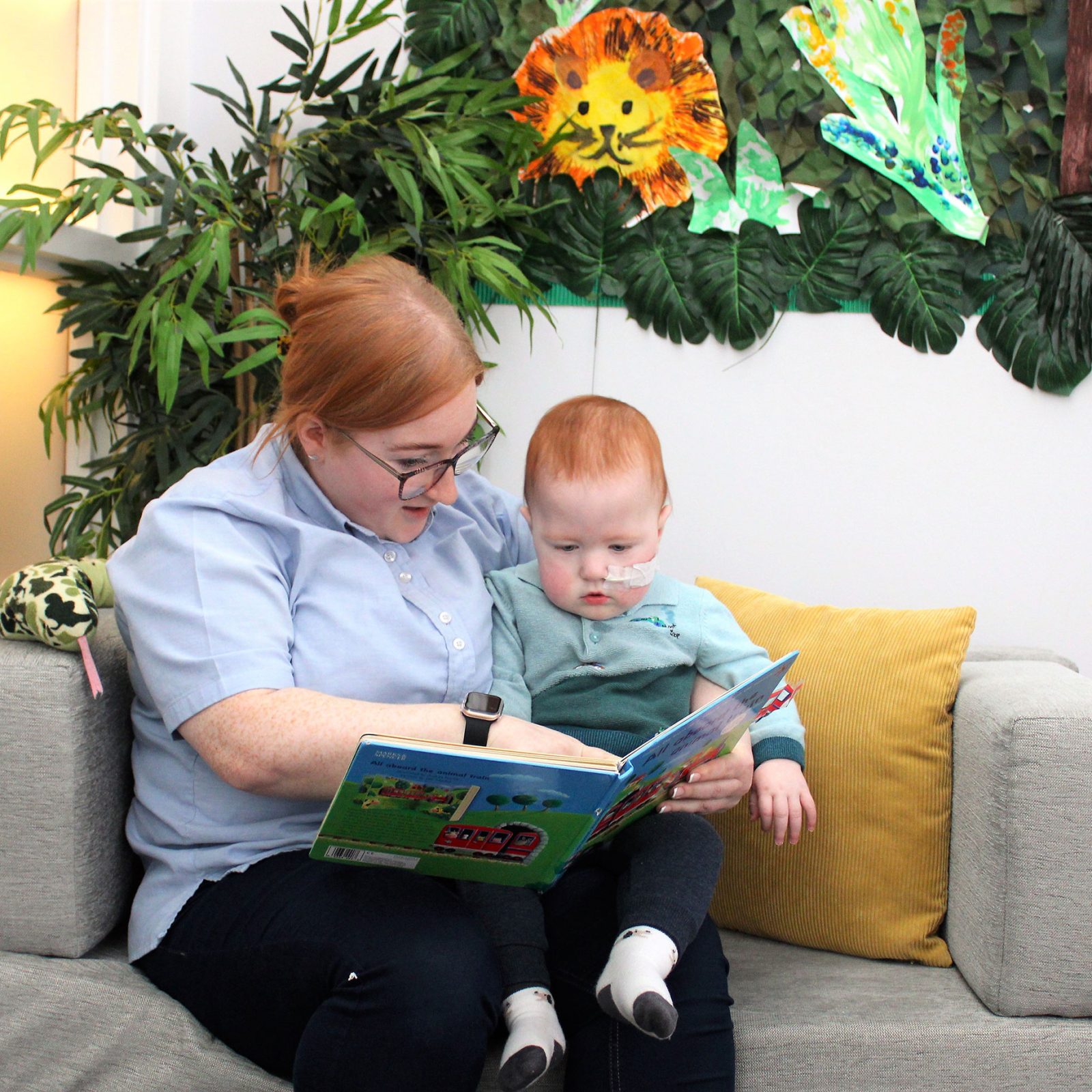 Acorns Nurseries A person reads a book to a child sitting on a couch, surrounded by jungle-themed wall decorations and scattered acorns, evoking the enchanting atmosphere of Parc Nantgarw.