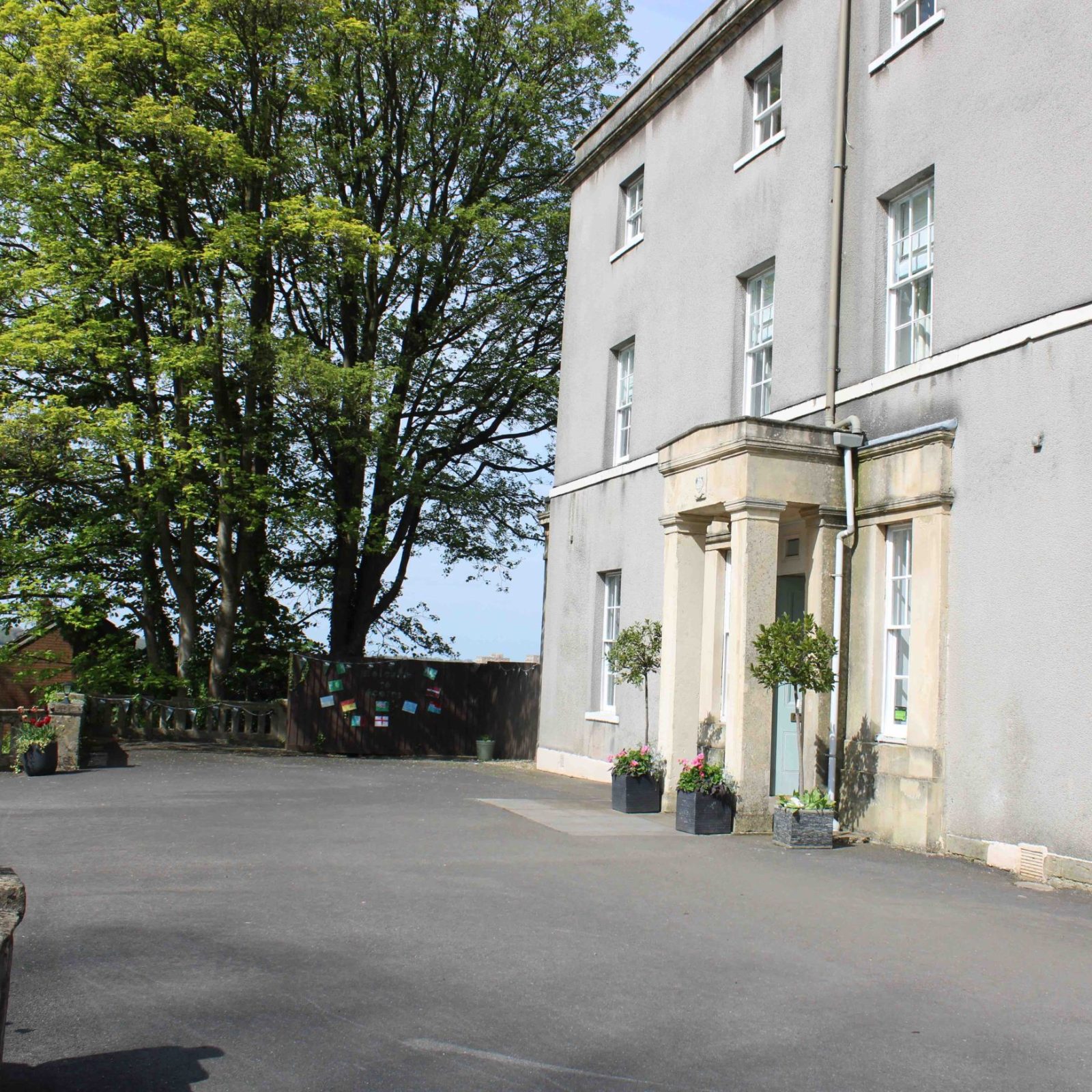 Acorns Nurseries The large, gray stone building, known as Henbury Hill House, boasts multiple windows and is gracefully surrounded by a paved area and an ancient tree with acorns gently scattered at its base.