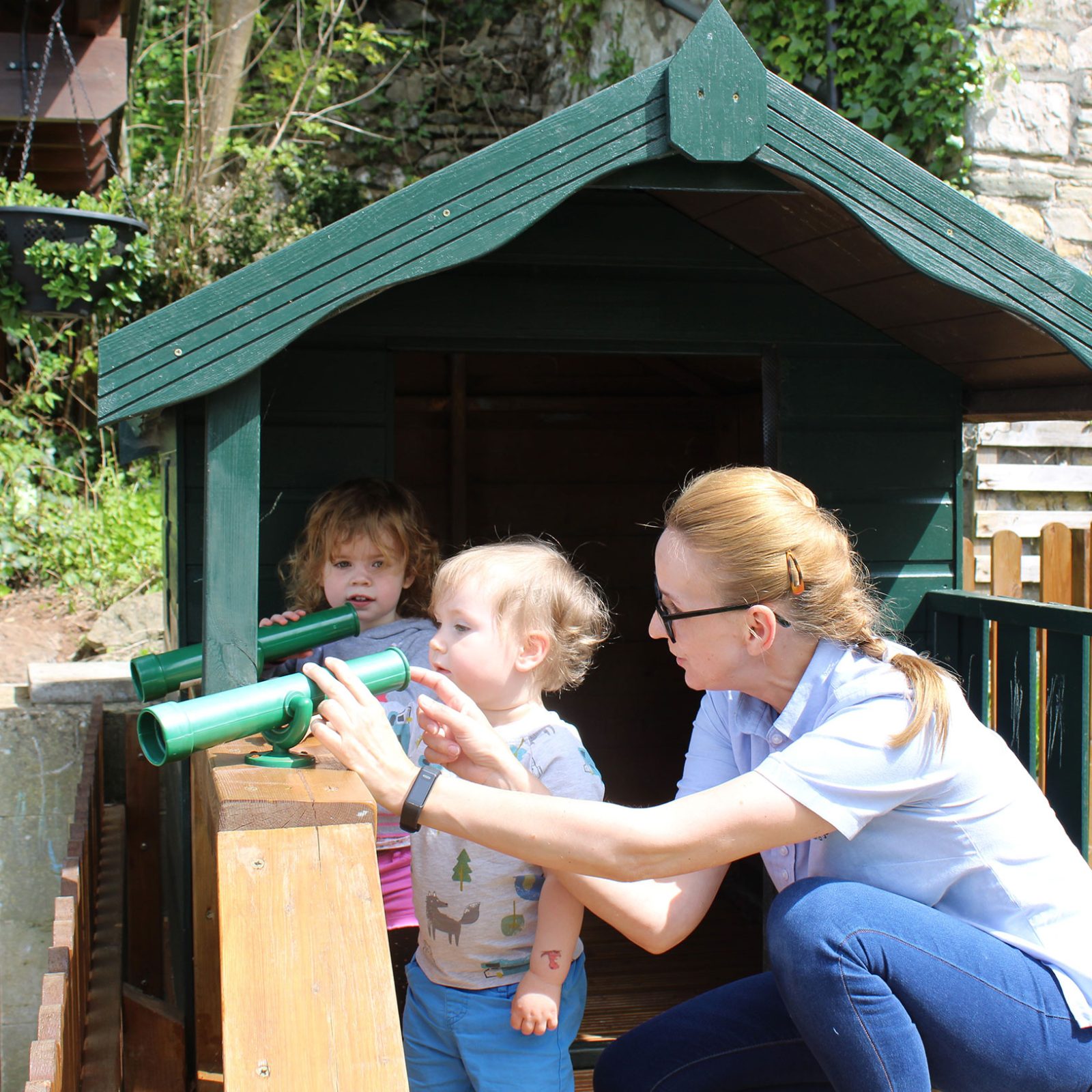 Acorns Nurseries At Henbury Hill House, an adult helps two children use toy binoculars at a small wooden playhouse, nestled among acorns in a charming garden setting.