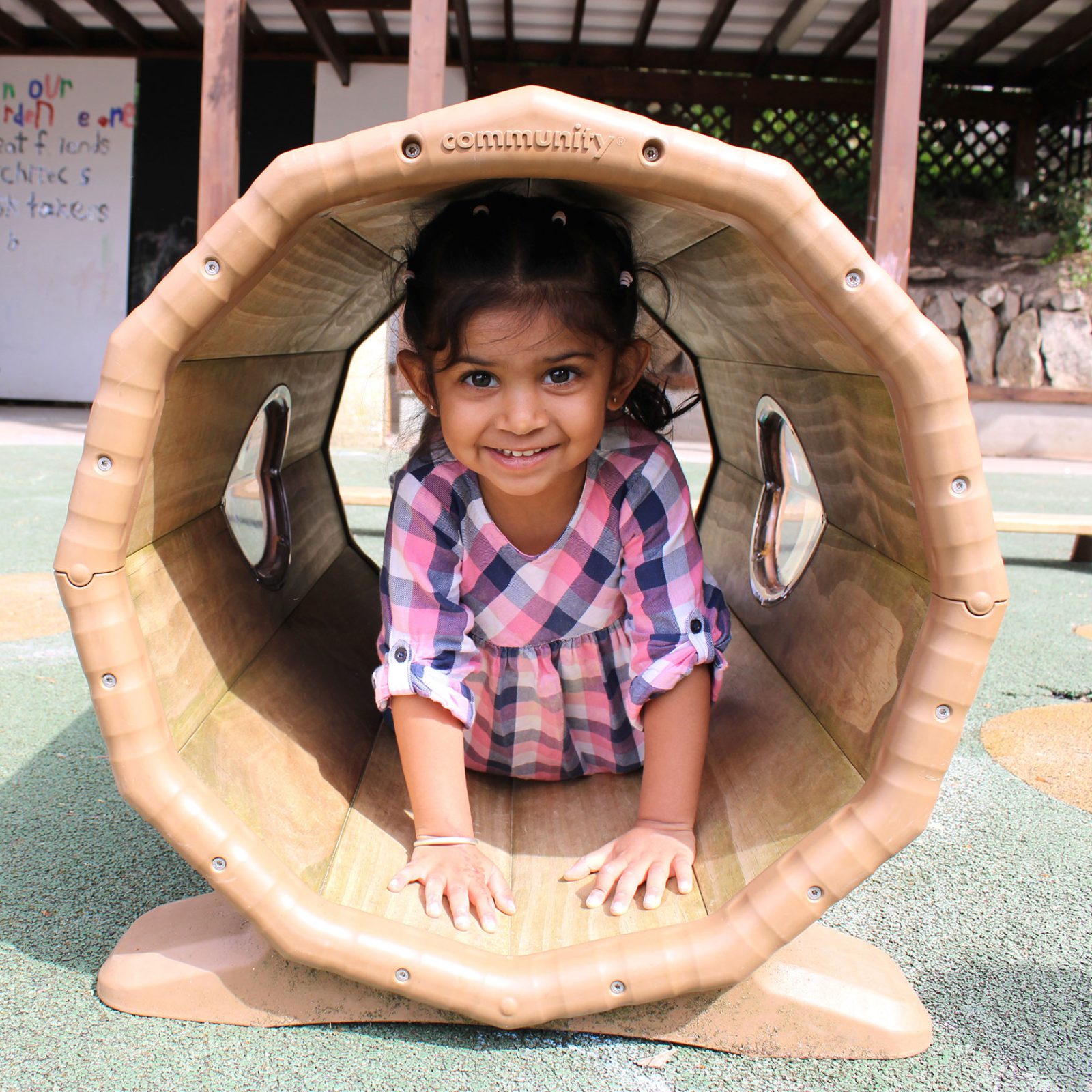 Acorns Nurseries A child in a plaid dress crawls through a wooden tunnel on the Henbury Hill playground, smiling at the camera, with scattered acorns adding an autumnal charm to the scene.