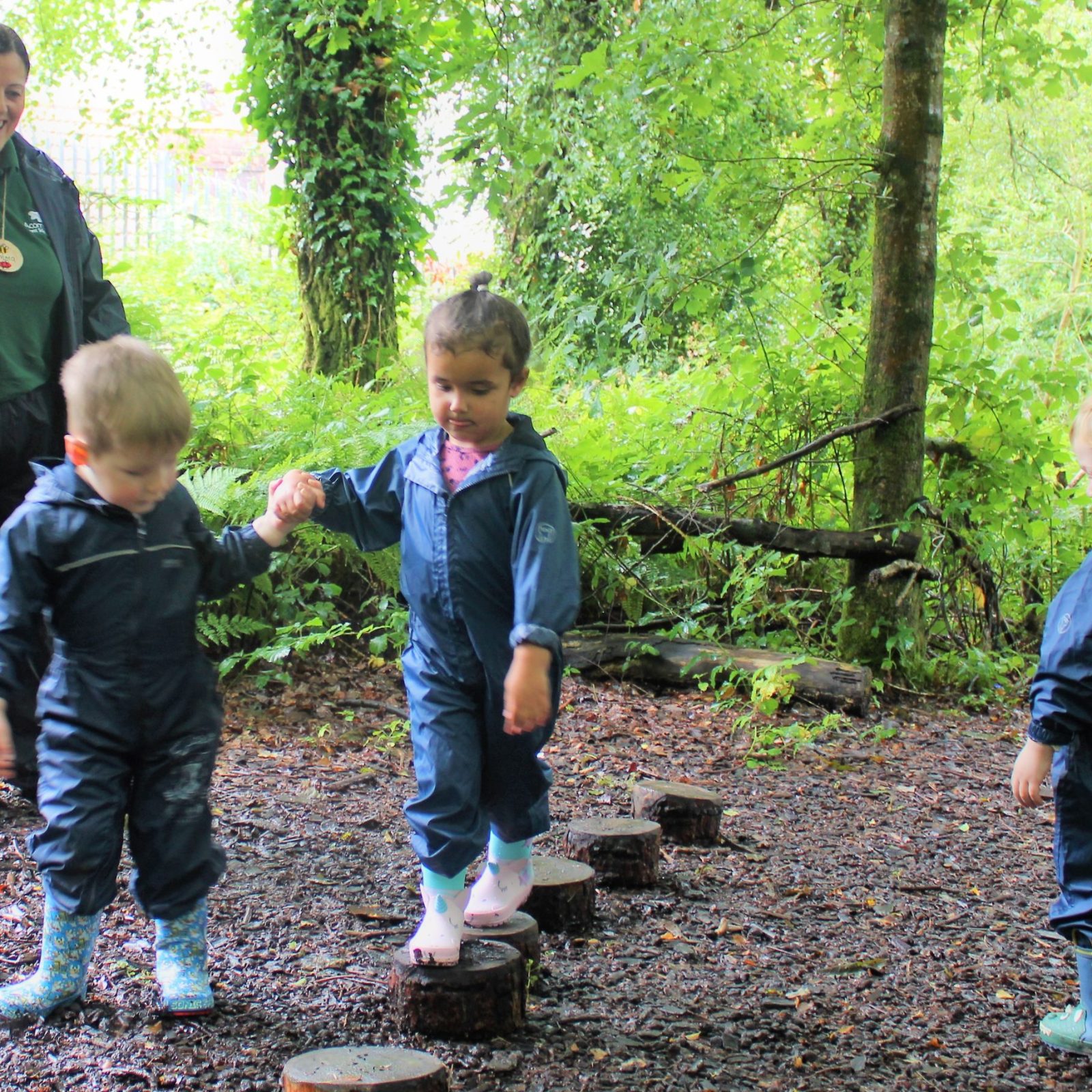 Acorns Nurseries Children wearing blue raincoats and boots balance on tree stumps in a forest, surrounded by scattered acorns, supervised by an adult in similar attire.