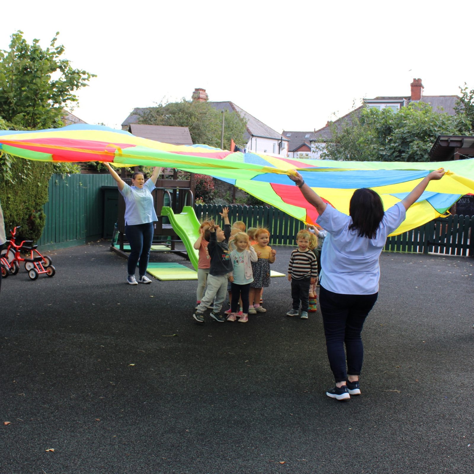 Acorns Nurseries Adults and children play with a colorful parachute in an outdoor playground area at Whitchurch, surrounded by trees, play equipment, and joyful moments resembling vibrant acorns.