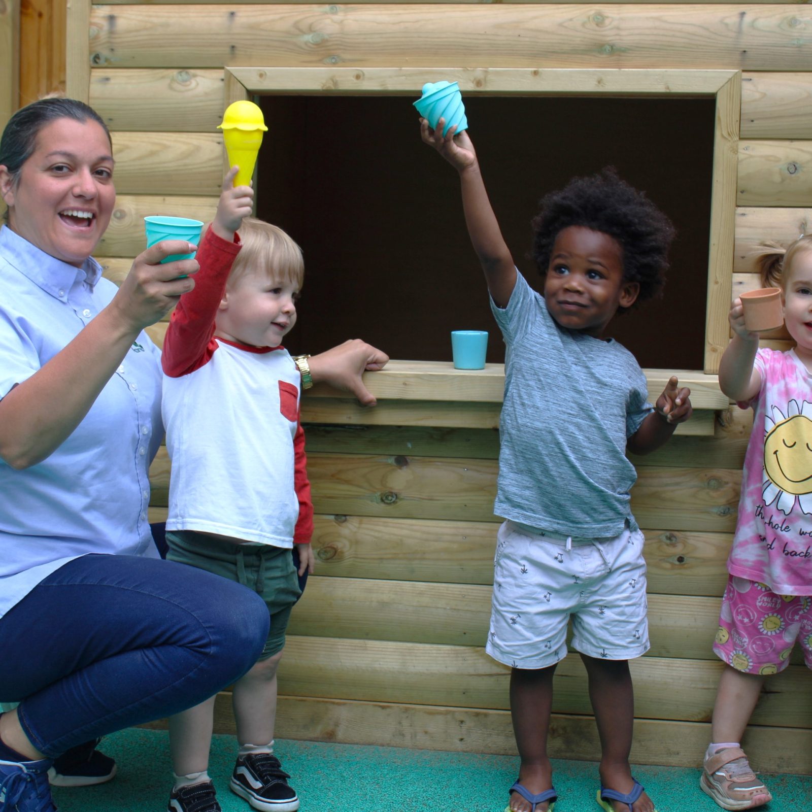 Acorns Nurseries Children and an adult hold up colorful ice cream cone toys in front of a wooden playhouse, filling the air with laughter at the Aviva workplace nursery.