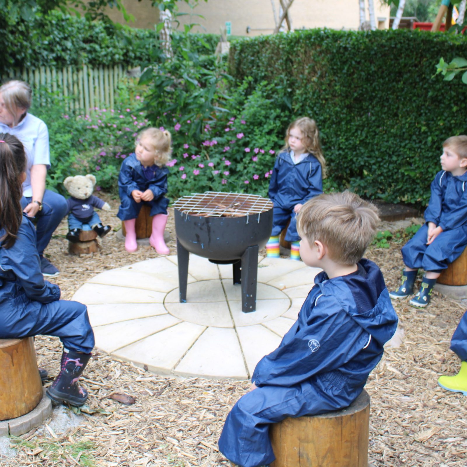 Acorns Nurseries Children in blue rain suits sit on wooden stumps in a circle outside at Aviva, listening to an adult with a book near a fire pit, much like a storytime session at a workplace nursery.