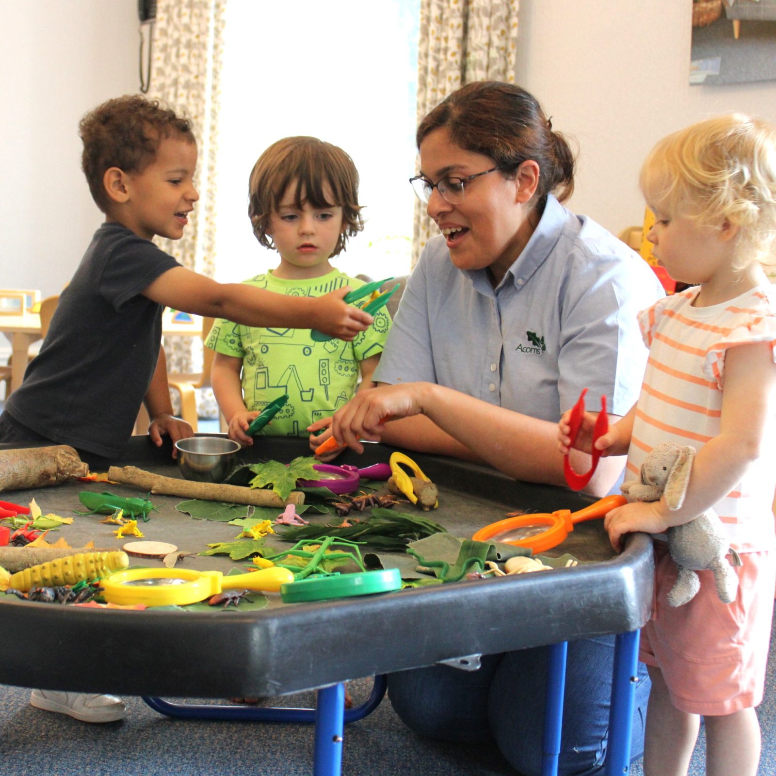 Acorns Nurseries At Henbury Hill House, children and a teacher engage in a nature-themed activity at a table adorned with leaves, sticks, acorns, and toy insects.