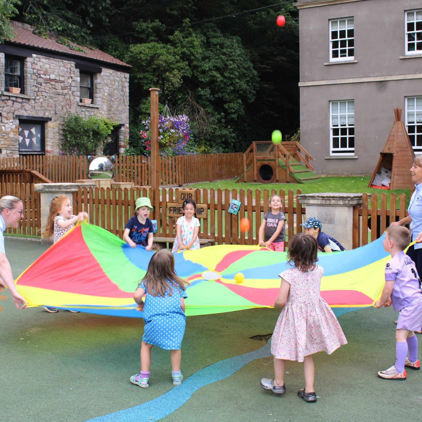 Acorns Nurseries Children and two adults joyfully play with a colorful parachute at the playground near Henbury Hill House, surrounded by trees and buildings, with acorns scattered across the ground.