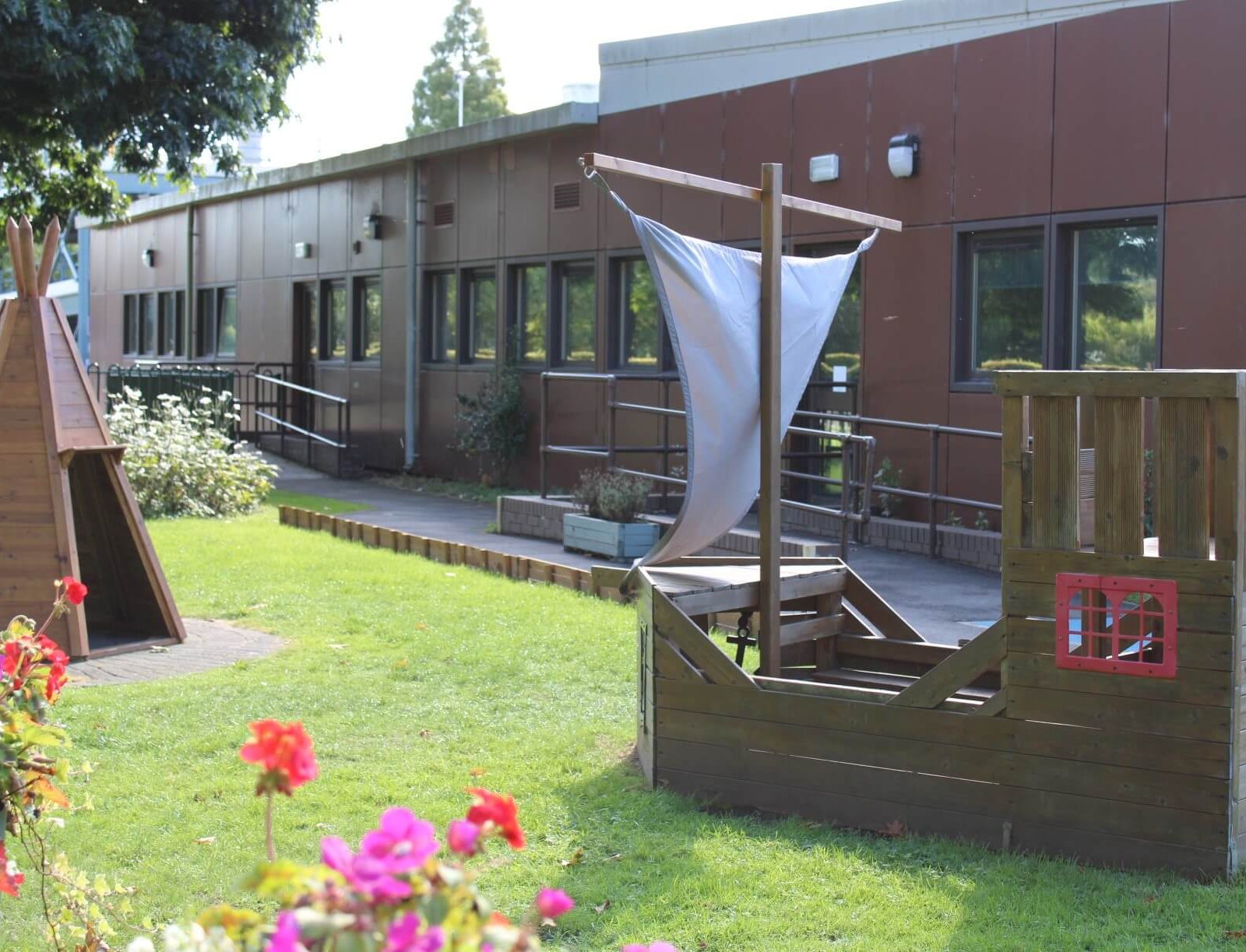 Acorns Nurseries Children's playground featuring a wooden pirate ship and a teepee structure on grass is conveniently located near the brown building, making it an ideal spot for an Acorns ONS Workplace Nursery.