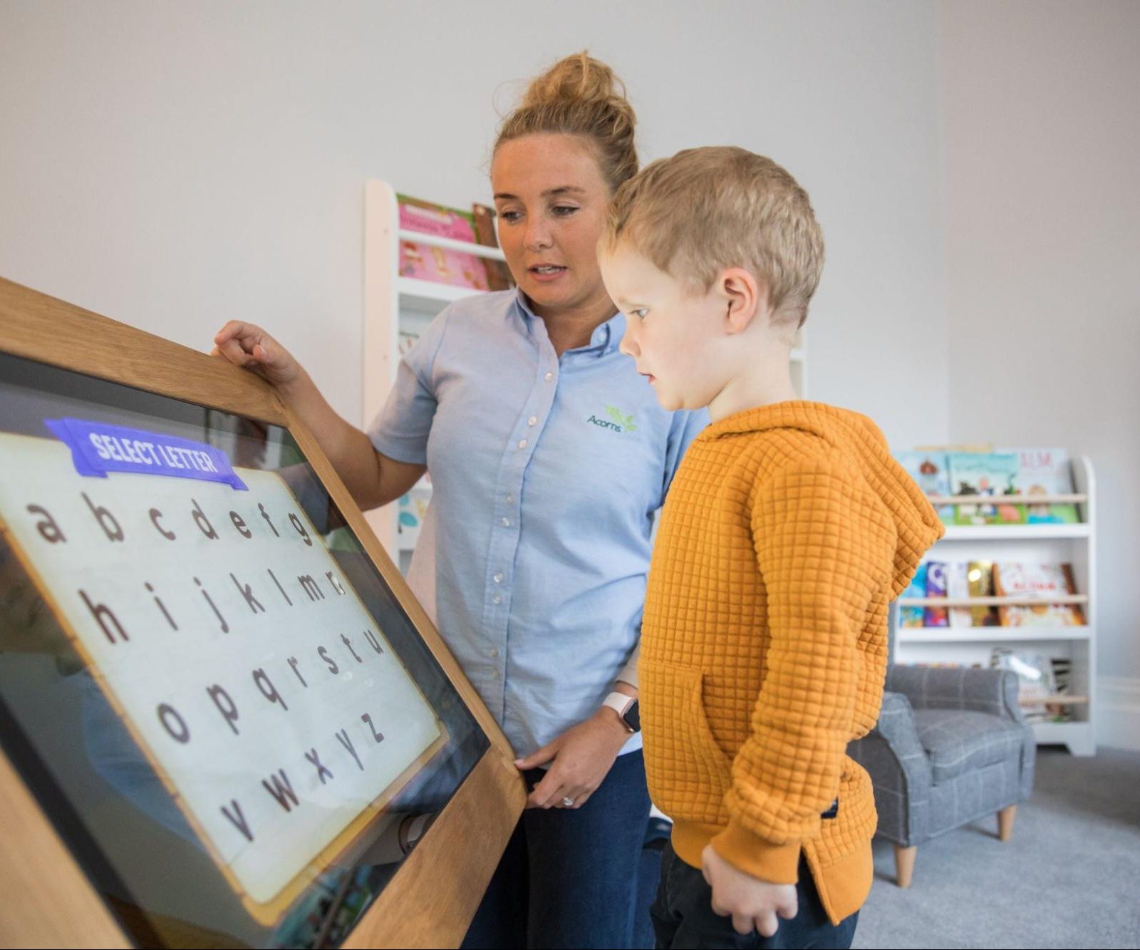 Acorns Nurseries A child explores an educational touchscreen, guided by an adult in a room filled with bookshelves and learning materials, choosing their path of discovery like planting acorns for future growth.