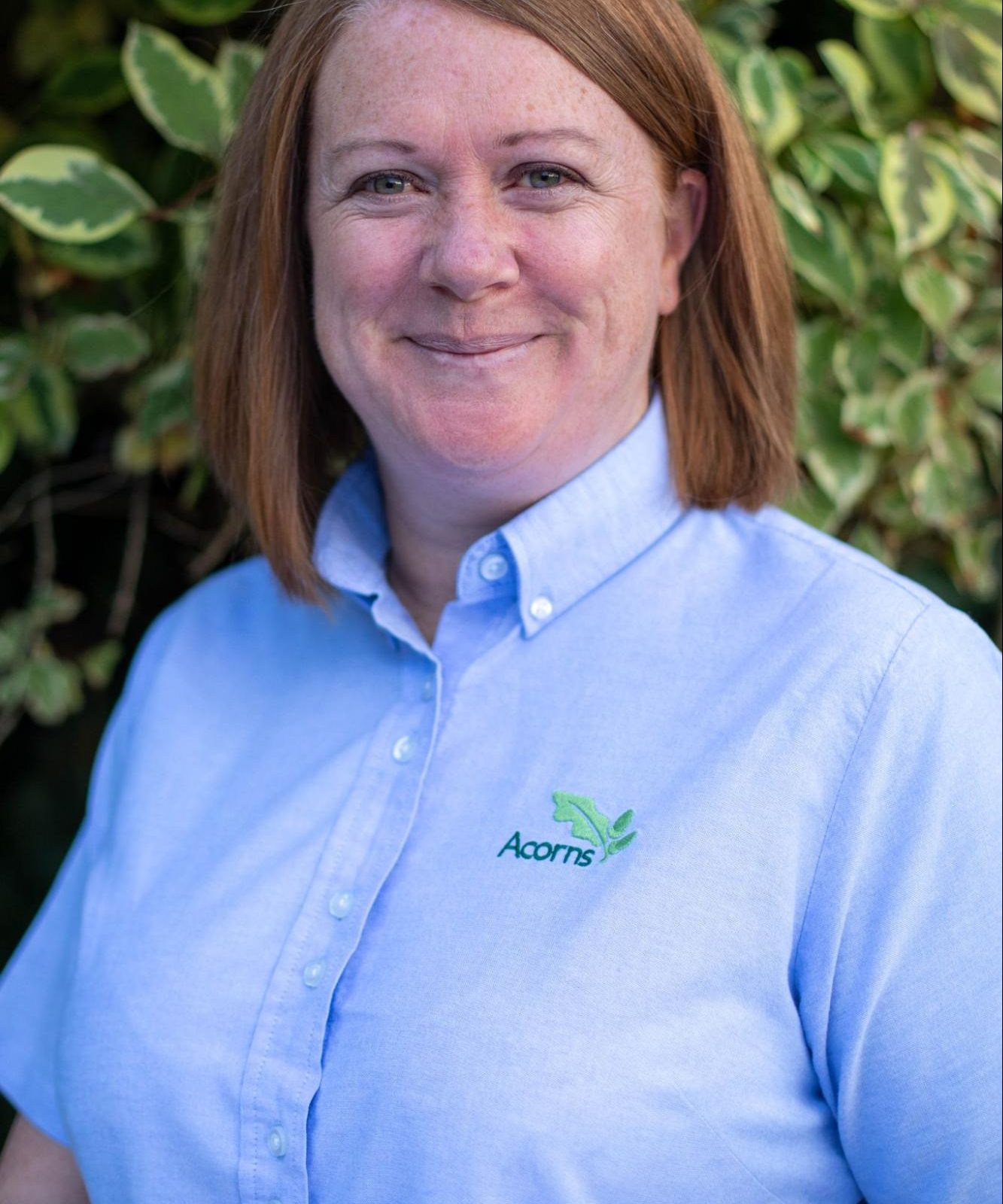 Acorns Nurseries A person with brown hair in a blue shirt stands in front of green foliage, smiling at the camera, while a charming house peeks through the trees.