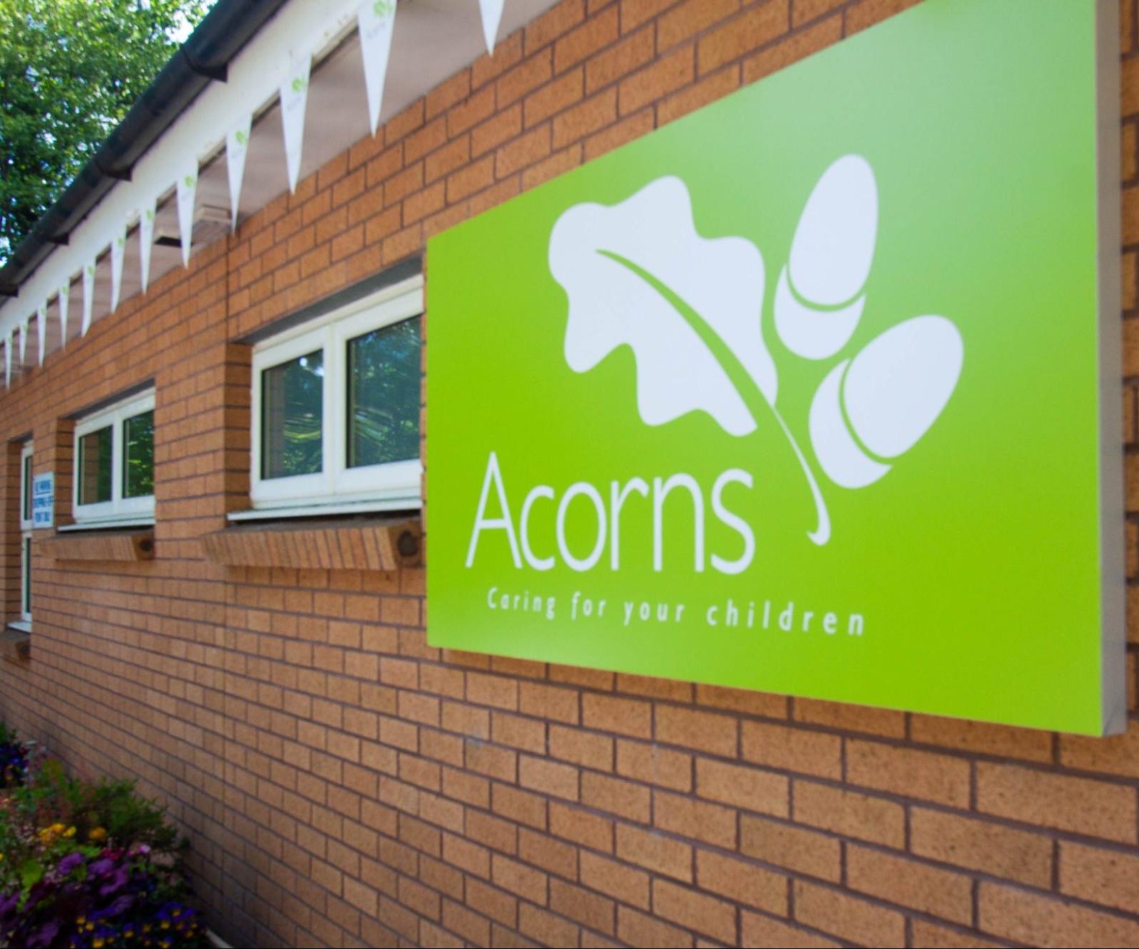 Acorns Nurseries A green sign with an acorn logo on a brick building reads, "Acorns Caring for Your Children." White flags hang above, creating a welcoming home-like atmosphere.