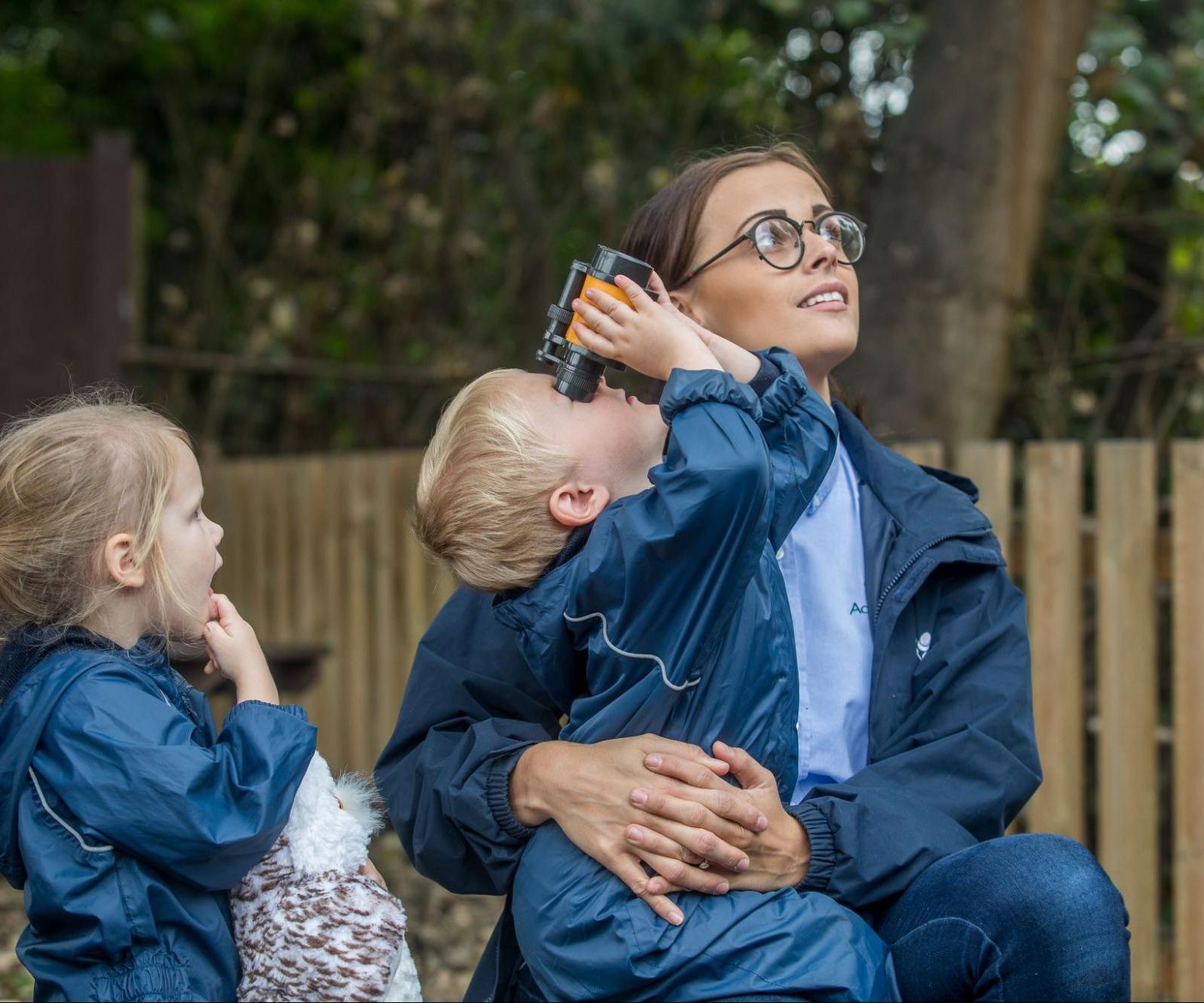 Acorns Nurseries A woman sits outdoors with her two children; the child on her lap looks through binoculars, while the other stands beside holding a toy owl. It's a perfect moment of exploration and learning, reminiscent of a day at Forest School.