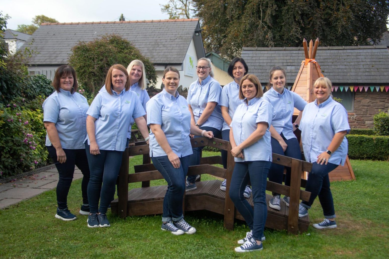 Acorns Nurseries Nine women, part of a dedicated team, in matching blue shirts and jeans stand and sit on a wooden structure in a garden setting with a tepee in the background.