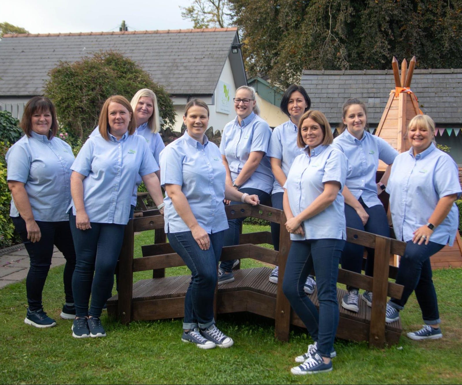 Acorns Nurseries Nine women, part of a dedicated team, in matching blue shirts and jeans stand and sit on a wooden structure in a garden setting with a tepee in the background.