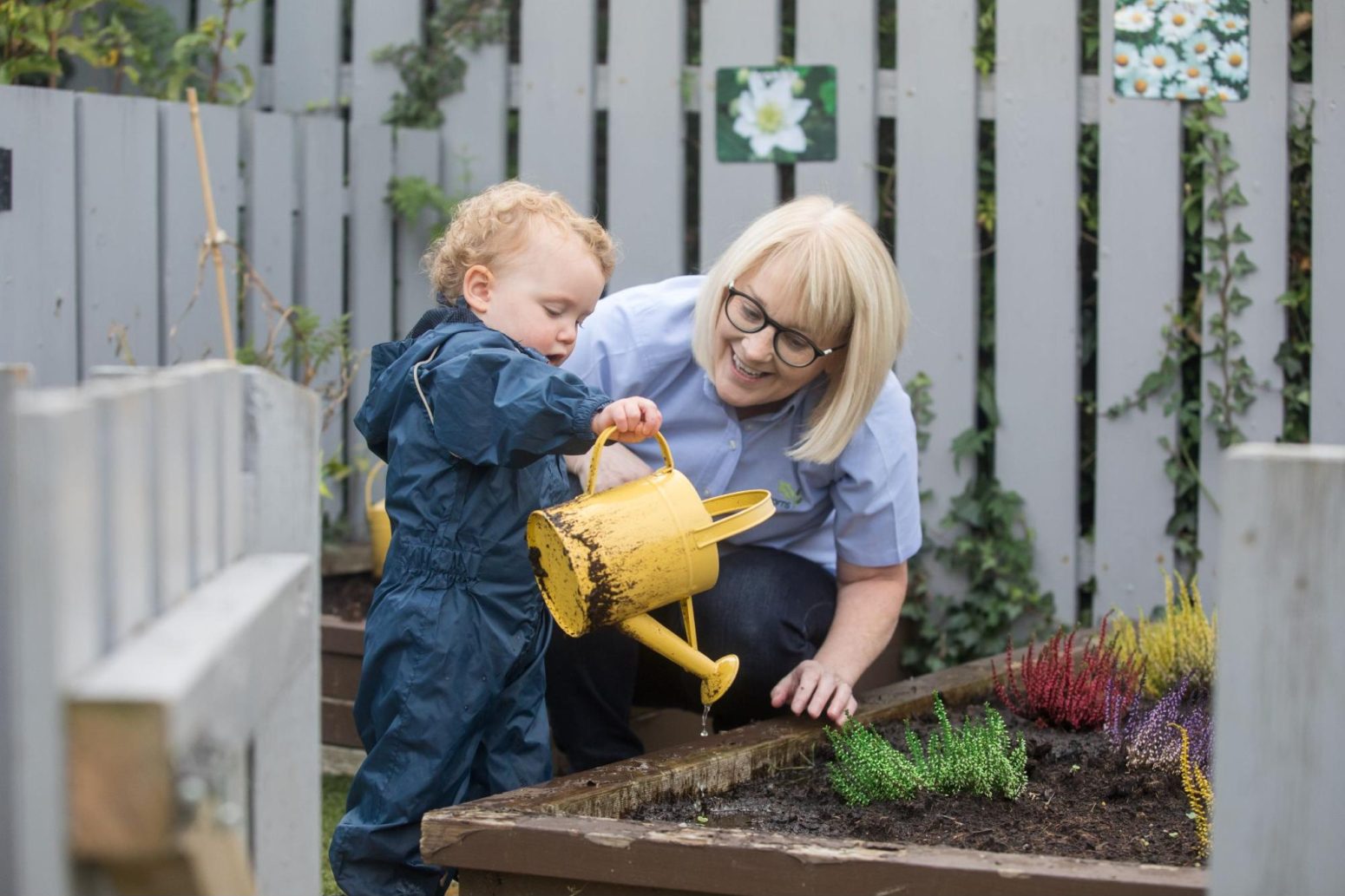 Acorns Nurseries A woman and a child in a garden share a beautiful partnership; the child is watering plants with a yellow watering can.