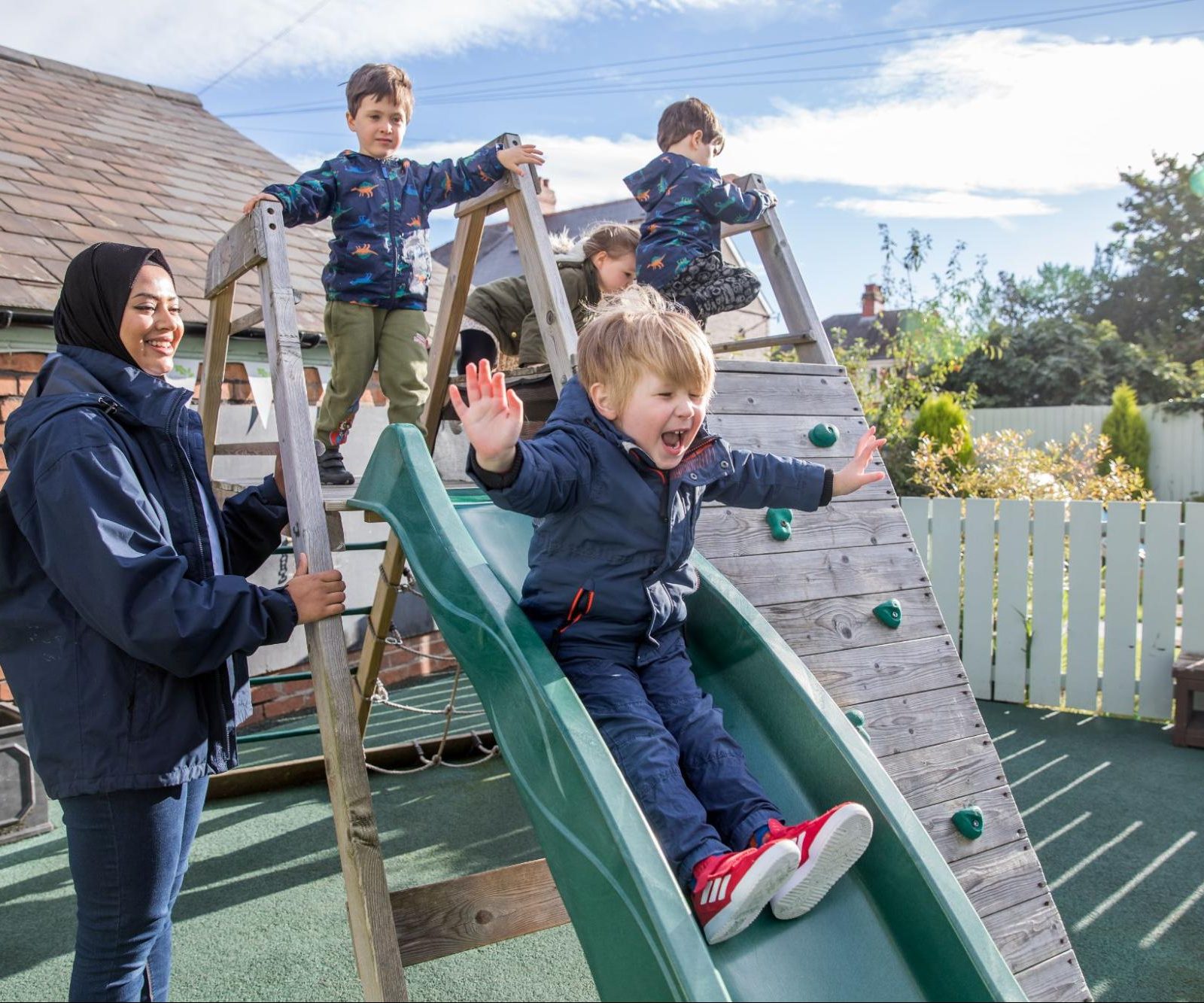 Acorns Nurseries Children enjoy the Great Outdoors as they play on a slide with an adult watching in the playground. One child joyfully slides down while others eagerly wait their turn.