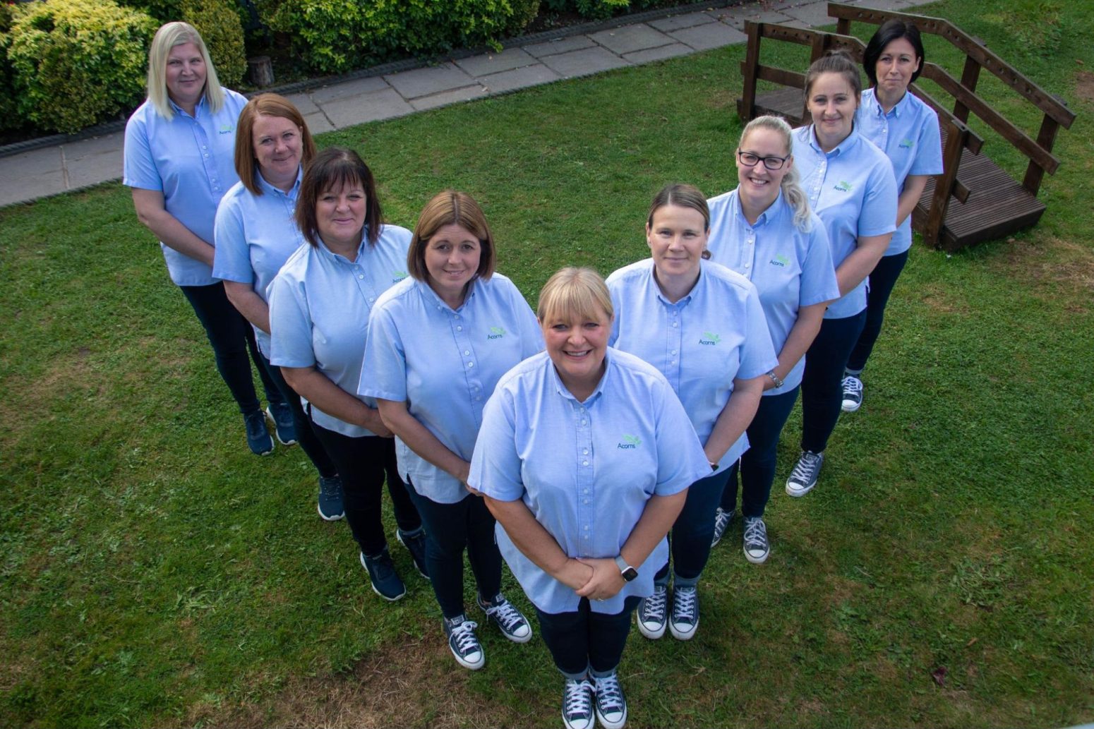 Acorns Nurseries A group of nine people wearing matching light blue shirts stands in a V-formation on a grassy area, embodying the spirit of home.