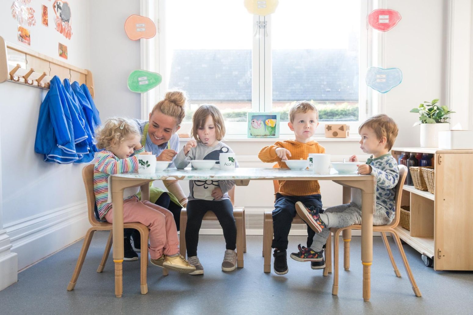 Acorns Nurseries Children are seated at a table in a classroom, enjoying healthy snacks under the watchful eye of an adult. The walls are adorned with colorful decorations, creating a cheerful atmosphere.