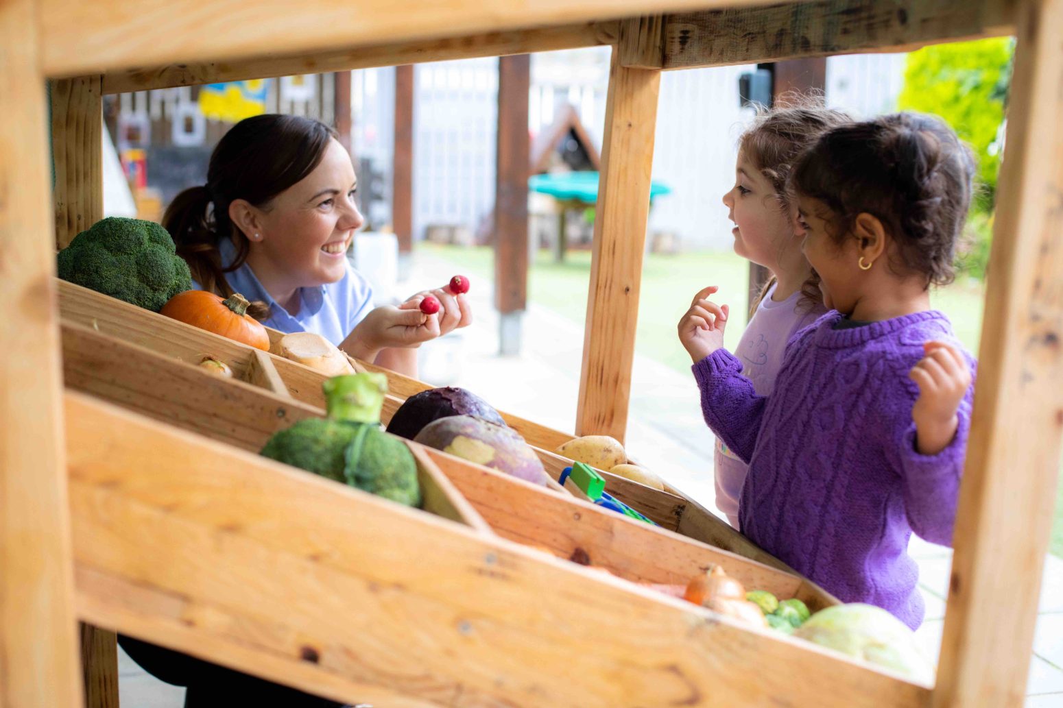 Acorns Nurseries A teacher and two children engage at a wooden vegetable stand, exploring the colorful assortment of veggies on display. Their conversation subtly weaves in lessons on investment, much like planting seeds for future abundance, echoing the Acorns philosophy of growth through smart beginnings.