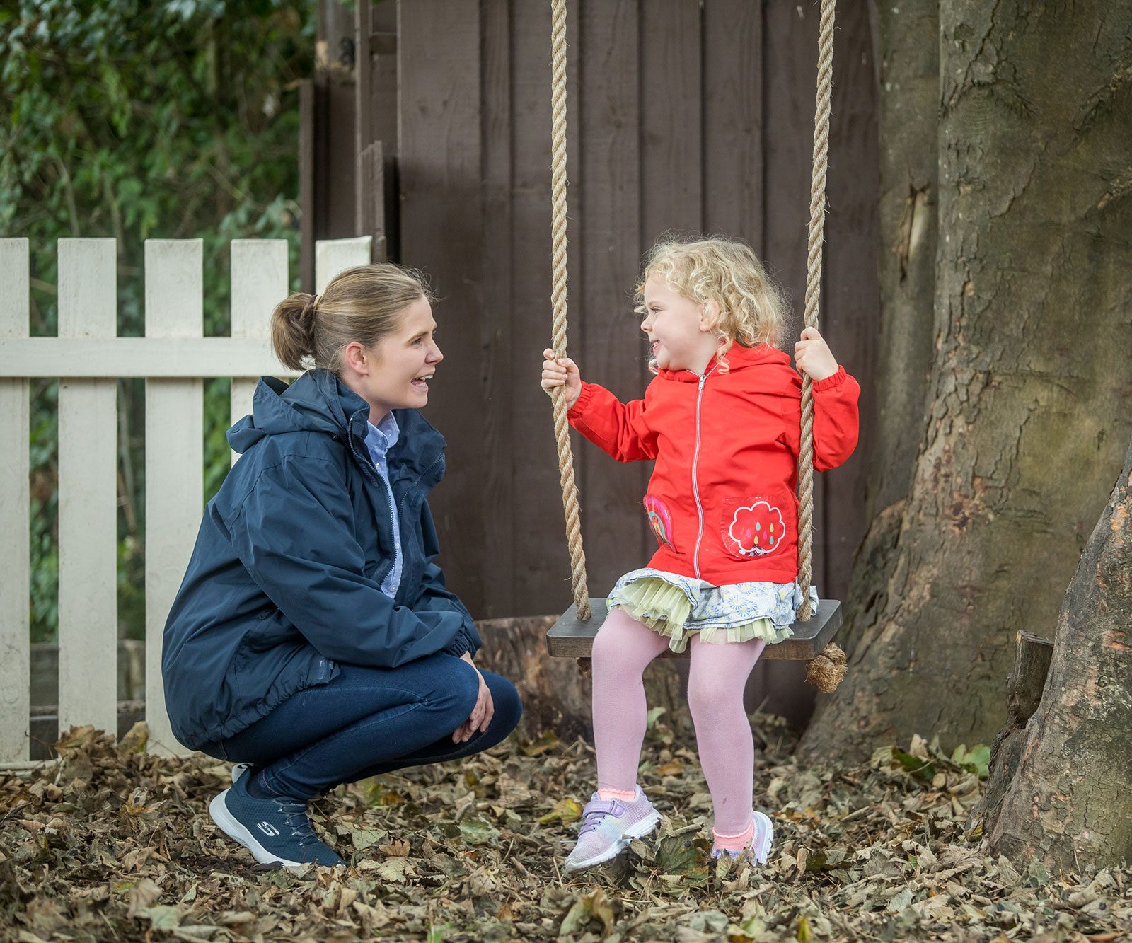 Acorns Nurseries An adult kneels beside a tree in Llanishen while a child in a red jacket sits on a swing, both smiling, surrounded by fallen leaves and scattered acorns.