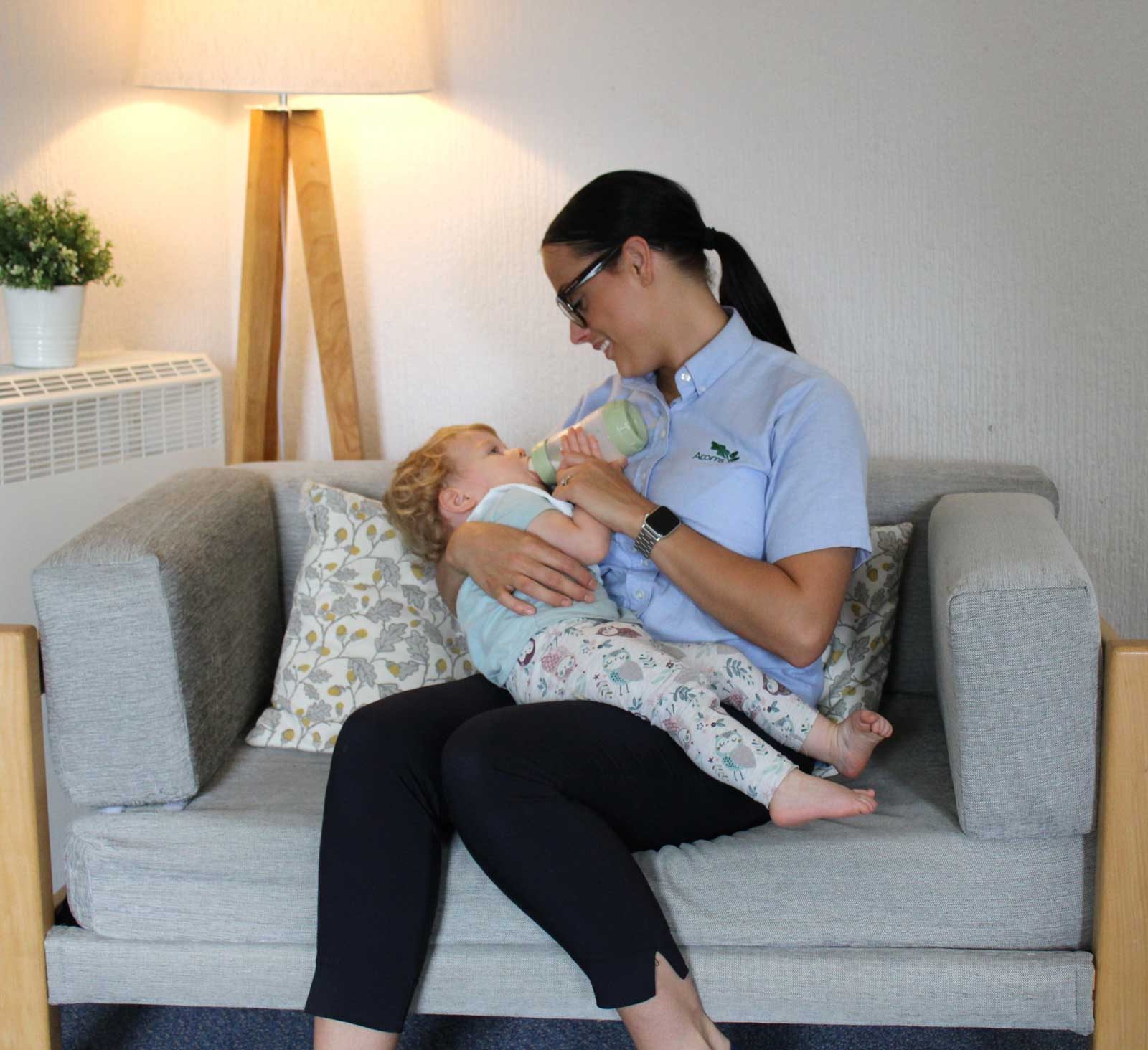 Acorns Nurseries A woman in Llanishen sits on a sofa, bottle-feeding a baby. A floor lamp and plant add a touch of warmth to the background, while acorns are scattered around like nature’s treasures.