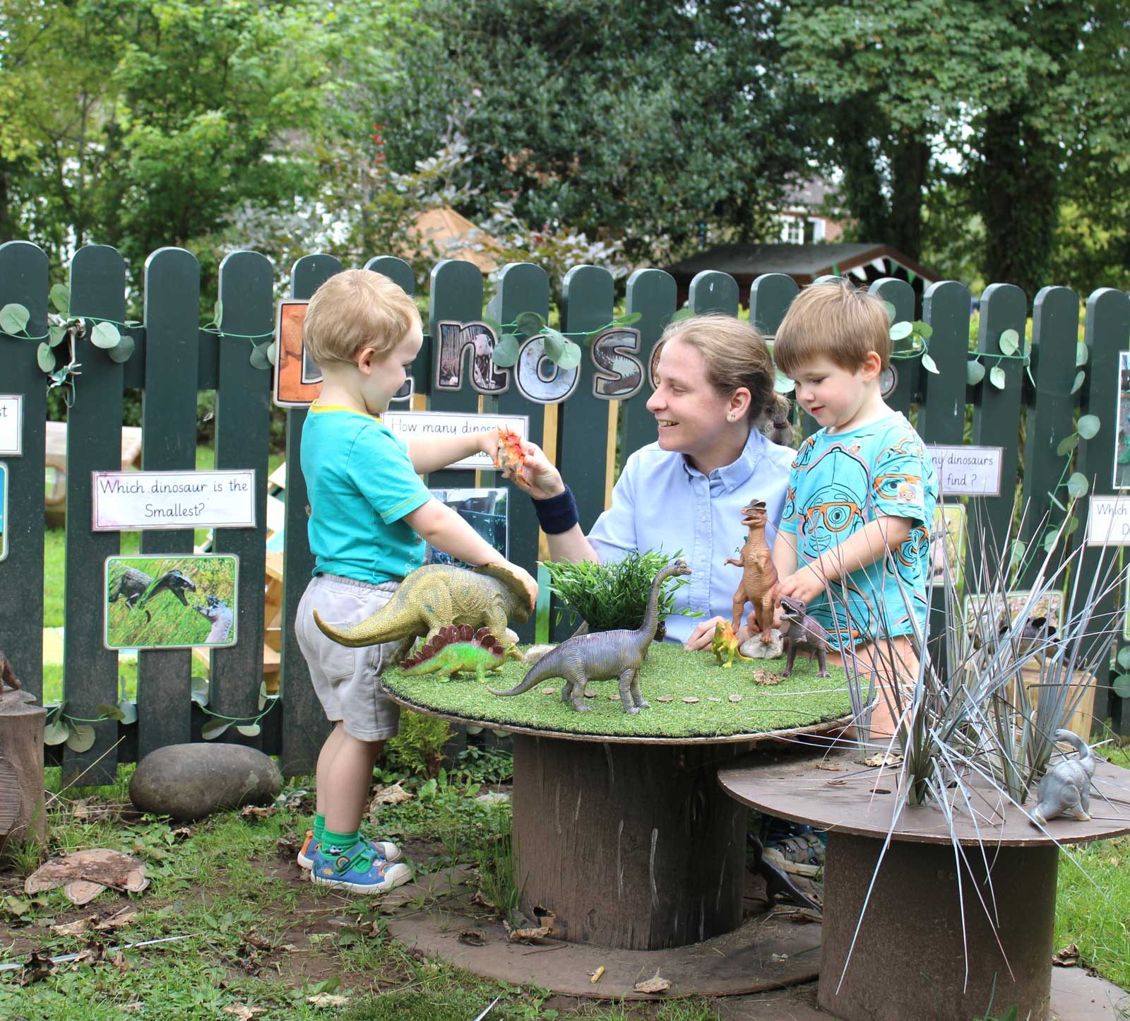 Acorns Nurseries At Acorns Llanishen, an adult and two children engage with dinosaur figurines on a table in an outdoor setting surrounded by informational signs and lush greenery.