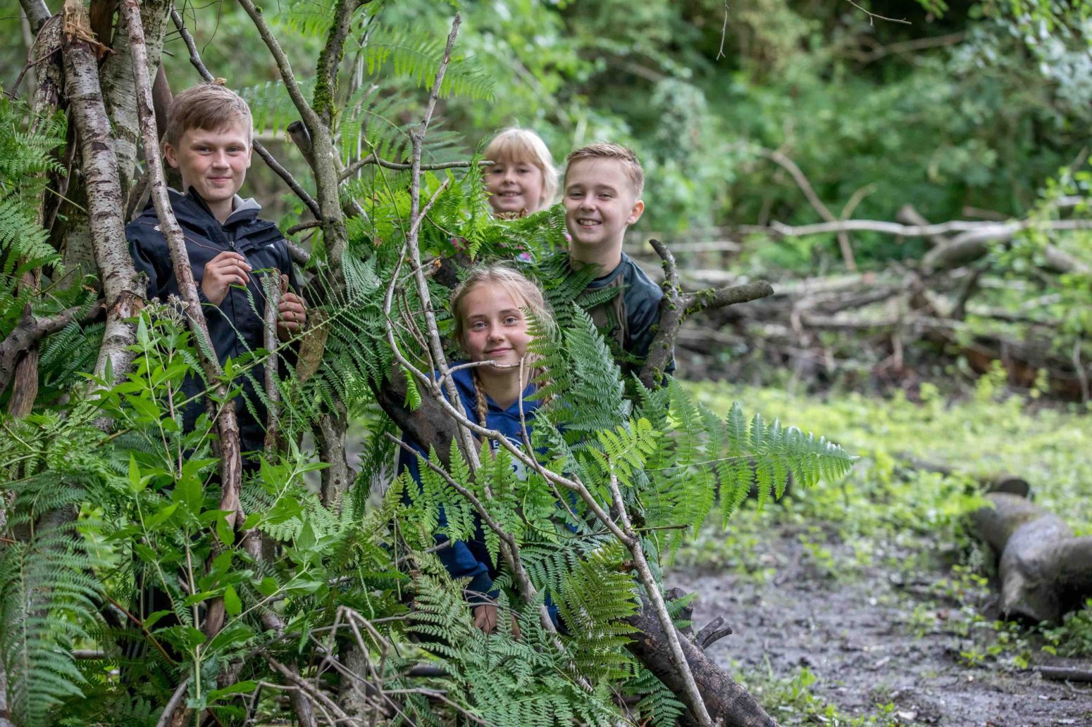 Acorns Nurseries Four children are building a shelter with branches and leaves in a forest setting, learning the true essence of teamwork and caring for each other amidst nature's embrace.