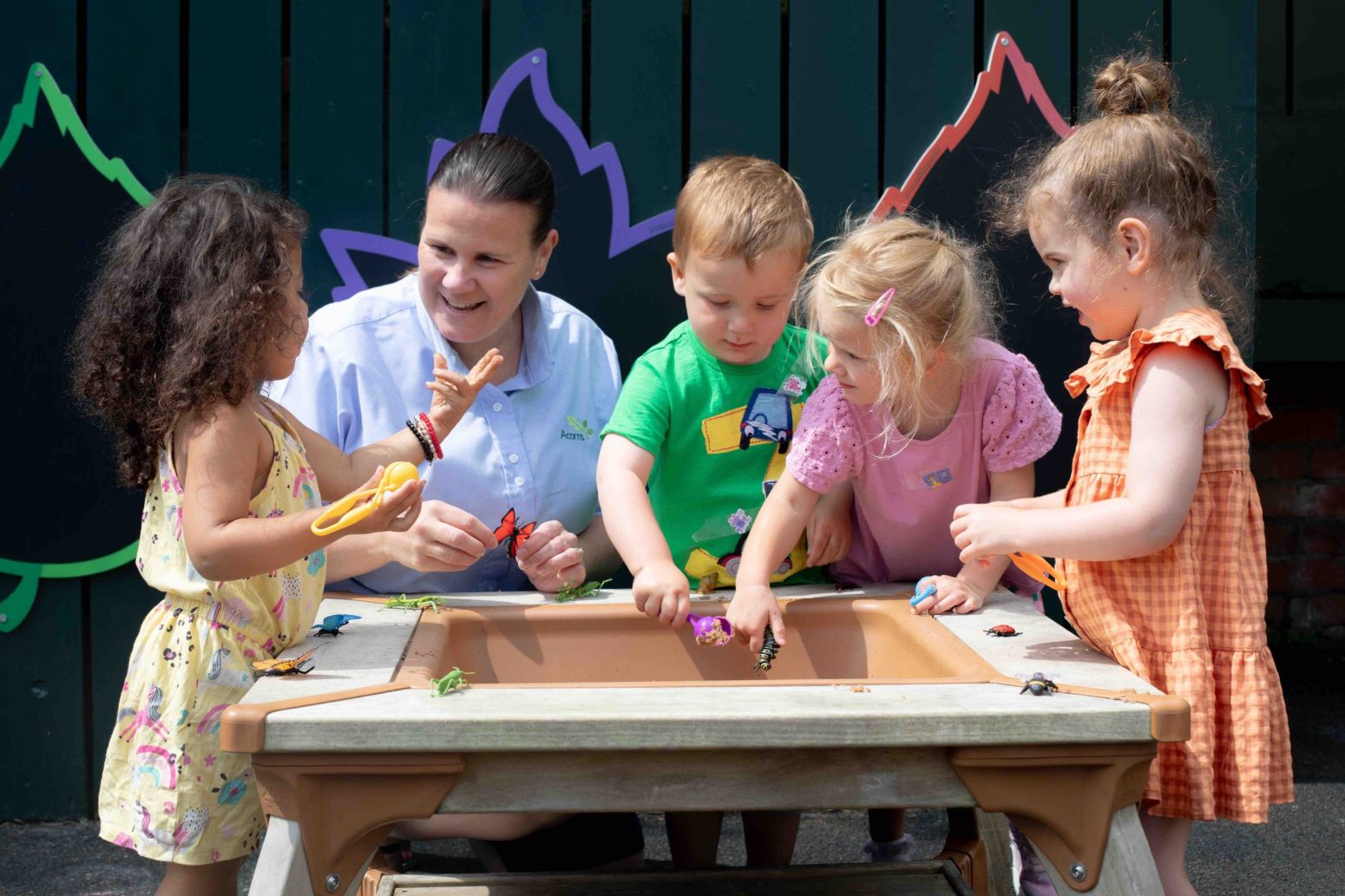 Acorns Nurseries Children and an adult engaging in playful education at a sand table, surrounded by colorful wall decorations outside.