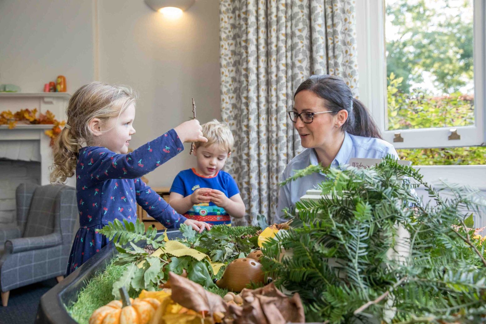 Acorns Nurseries Children and a caregiver engage with nature-themed objects at a table inside a room, where the large window provides an educational view that fosters care for the environment.