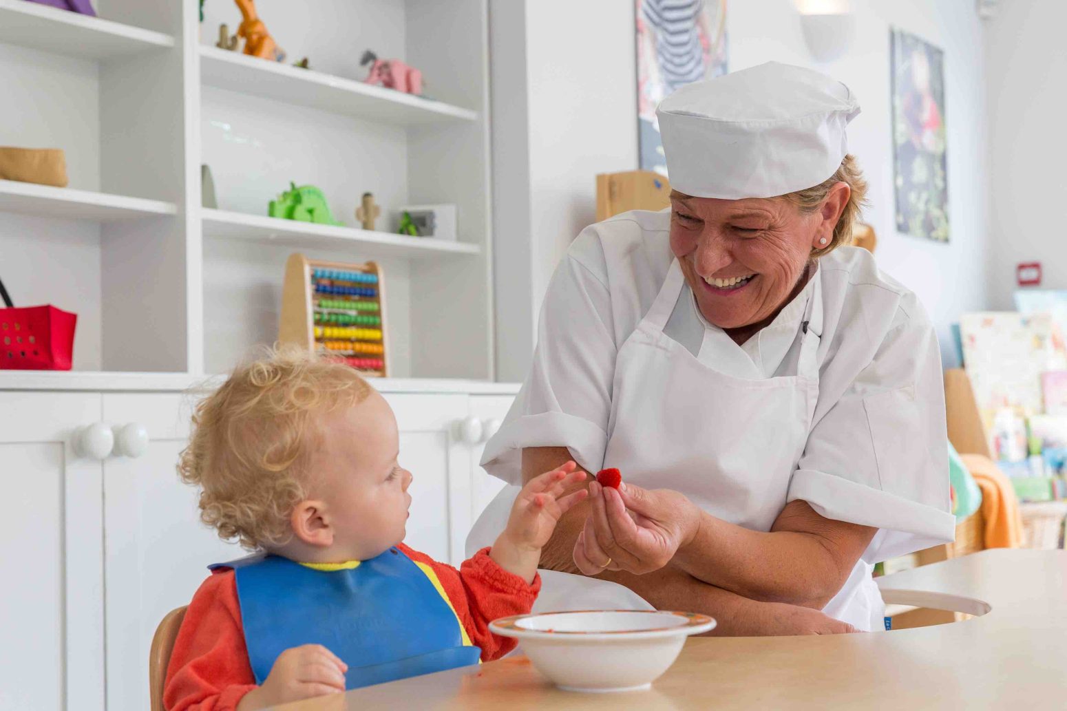 Acorns Nurseries A chef in a white uniform, embodying the spirit of teamwork, smiles at a young child as they hand them a red fruit. In the background, shelves hint at the collaborative environment of the bustling kitchen.