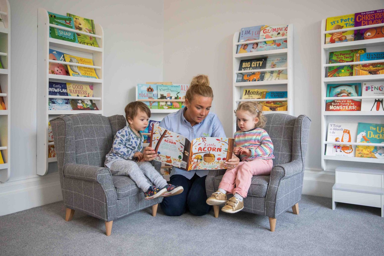 Acorns Nurseries An adult educates two children by reading a picture book to them as they sit on armchairs in a cozy room lined with bookshelves.