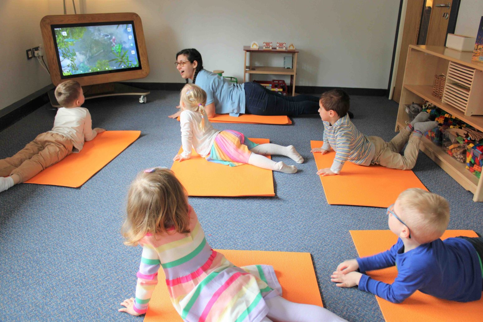 Acorns Nurseries Children and an adult sit on orange mats, learning the importance of teamwork as they watch an educational program on a screen in a classroom setting.