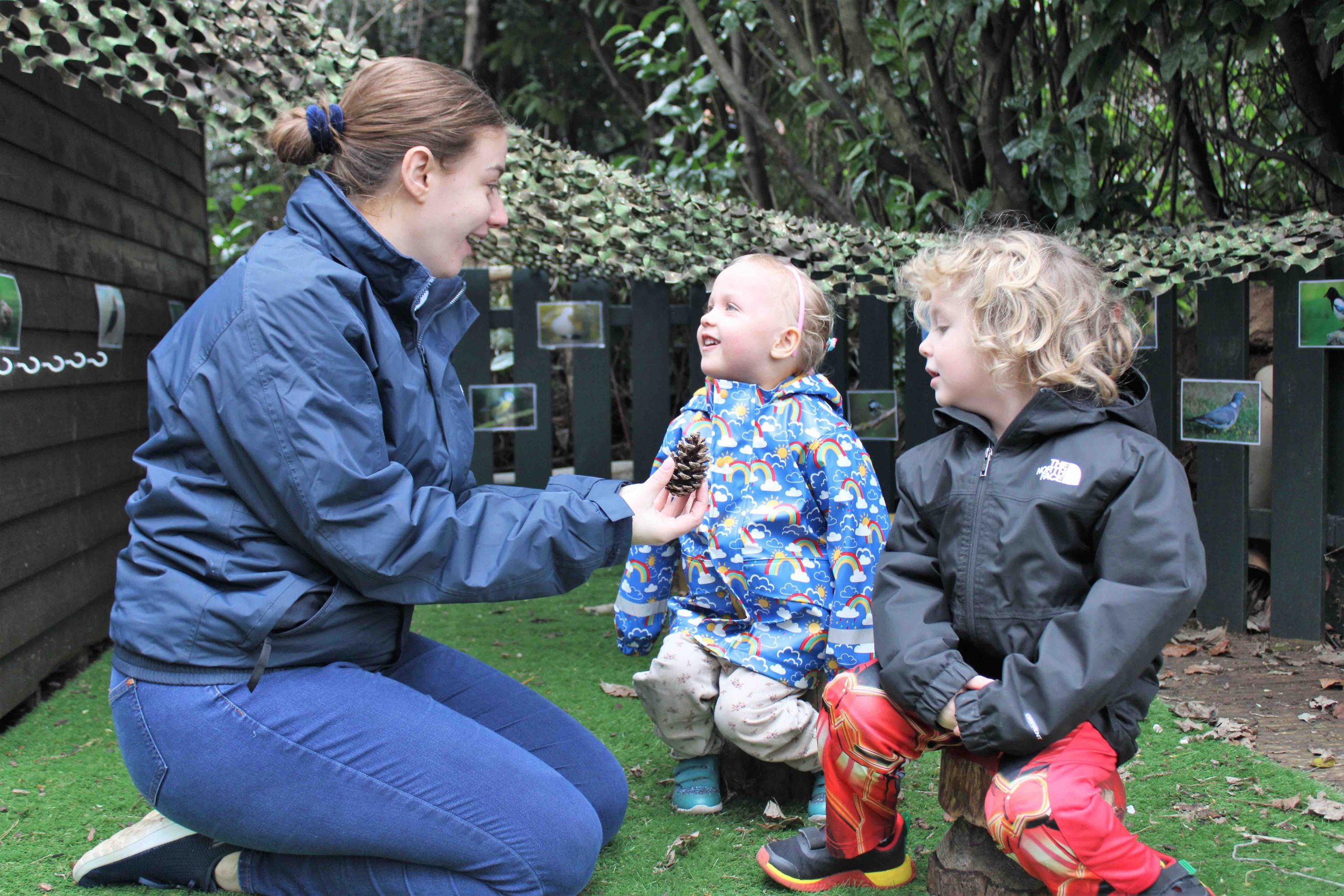 Acorns Nurseries A woman educating two young children outdoors shows them a pine cone amidst the lush greenery, creating an inspiring backdrop for discovery.