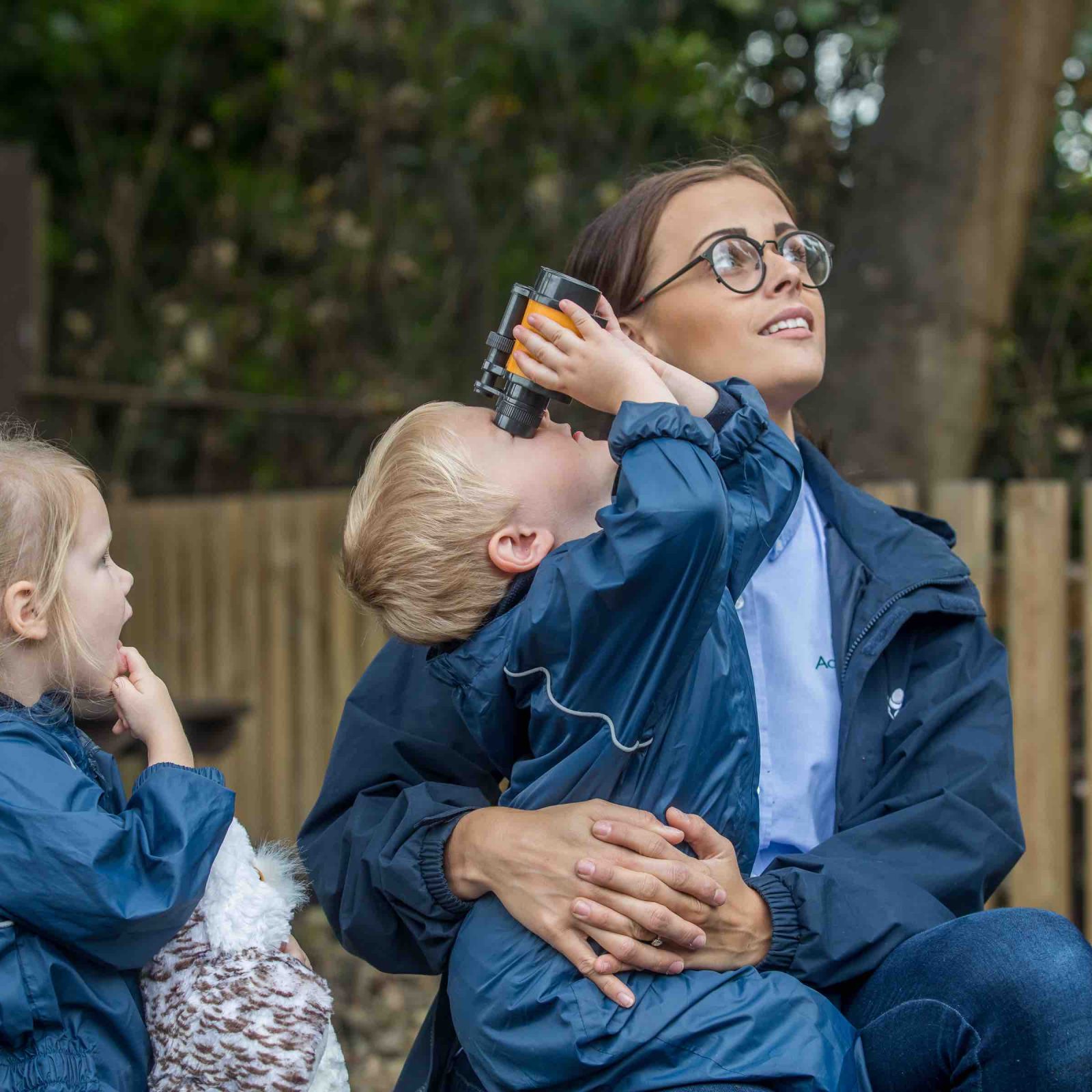 Acorns Nurseries A woman and two children, clad in cozy jackets, enjoy a home-away-from-home experience outdoors. One child eagerly peers through binoculars, capturing the essence of adventure while staying warmly connected to their surroundings.