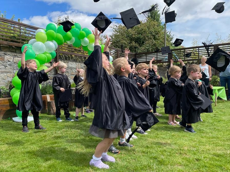 Acorns Nurseries Caring children wearing graduation gowns and caps celebrate joyfully outside, tossing their caps in the air next to vibrant green balloon decorations.