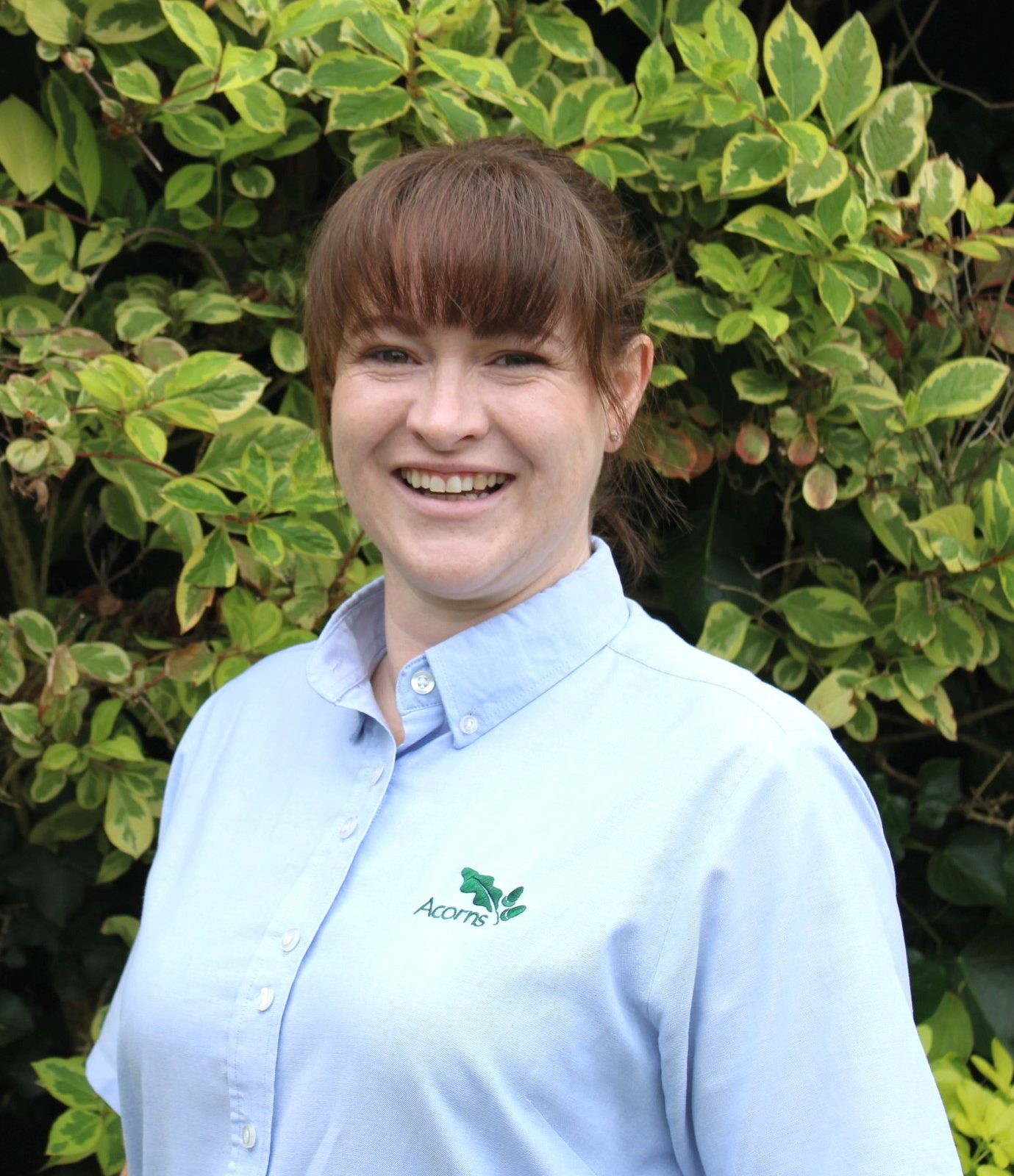 Acorns Nurseries A person wearing a blue shirt with an embroidered logo, possibly representing Llanishen, stands smiling in front of green foliage dotted with acorns.