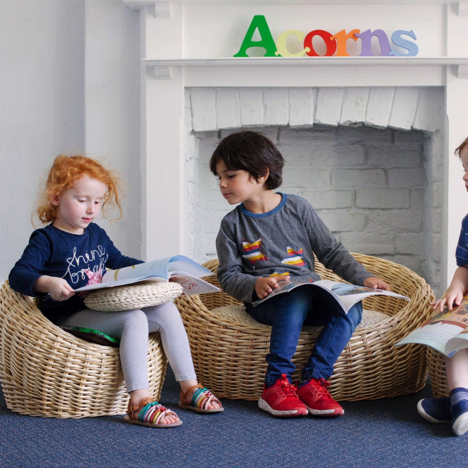 Acorns Nurseries Three children sit on wicker chairs in front of a cozy fireplace, reading books. The mantle displays block letters spelling 