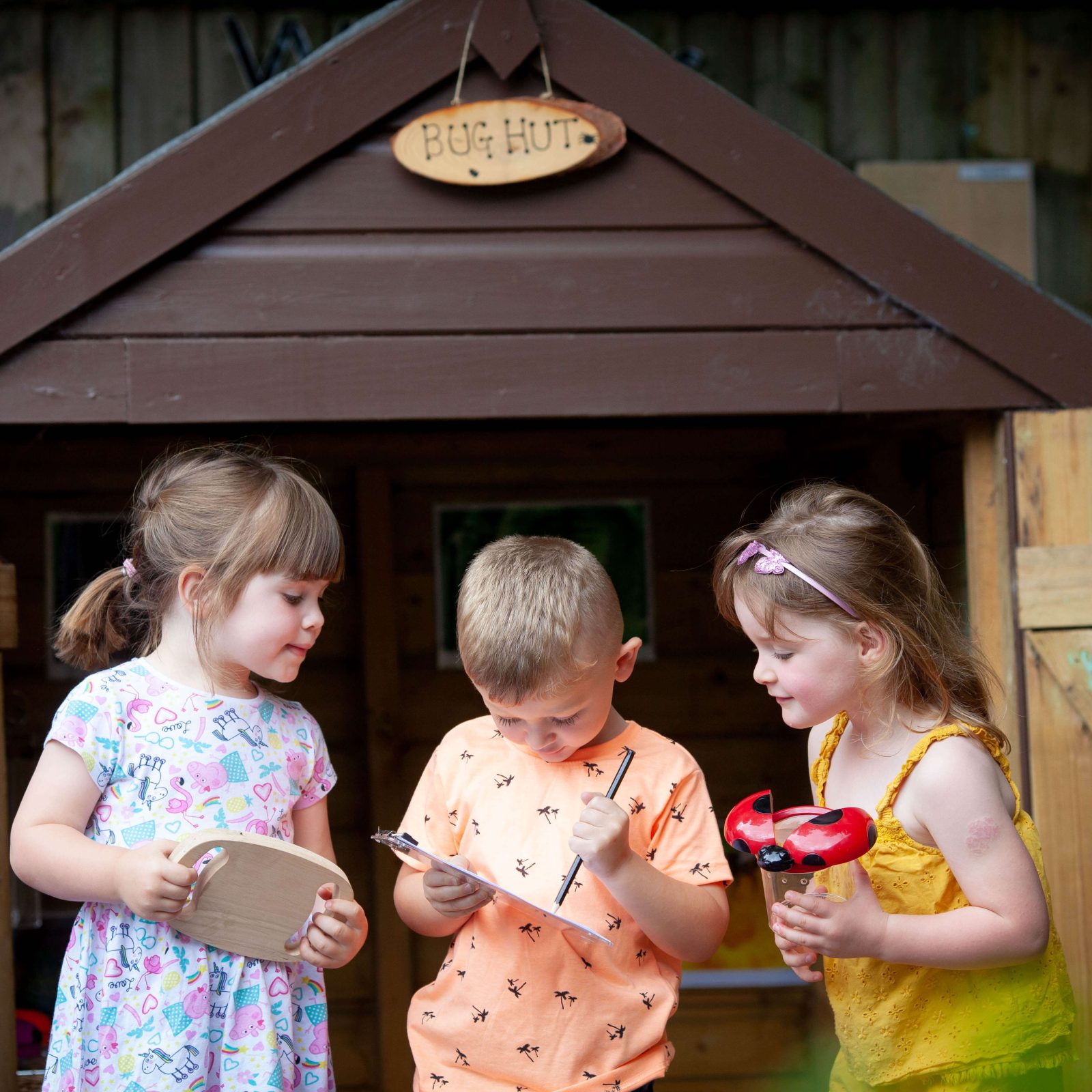 Acorns Nurseries Three children are standing outside a wooden playhouse that feels just like home, holding bug-catching equipment and a clipboard.