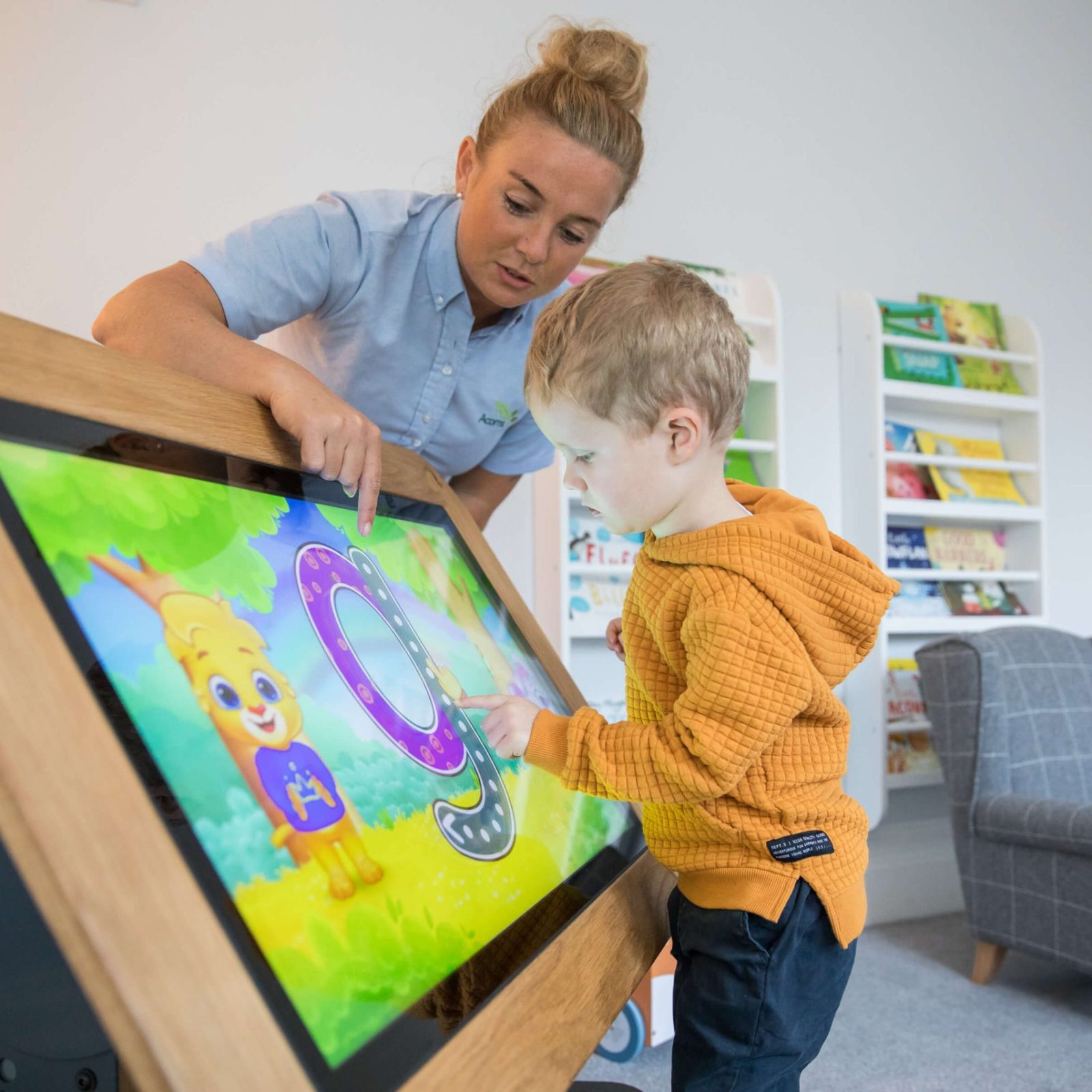 Acorns Nurseries A child interacts with a large touchscreen displaying an educational game, guided by an adult in a cozy home room lined with book shelves.