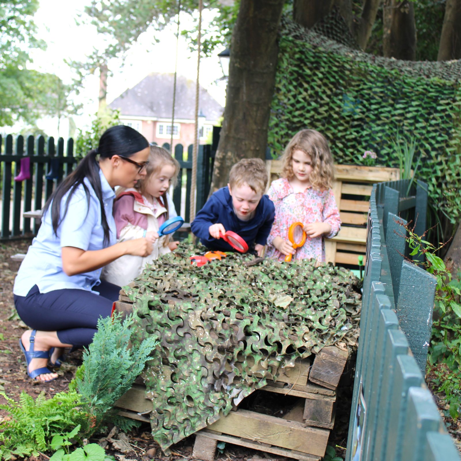 Acorns Nurseries A woman and three children explore a camouflaged outdoor area with magnifying glasses, near a fence and trees, searching for hidden acorns in Llanishen's lush landscape.