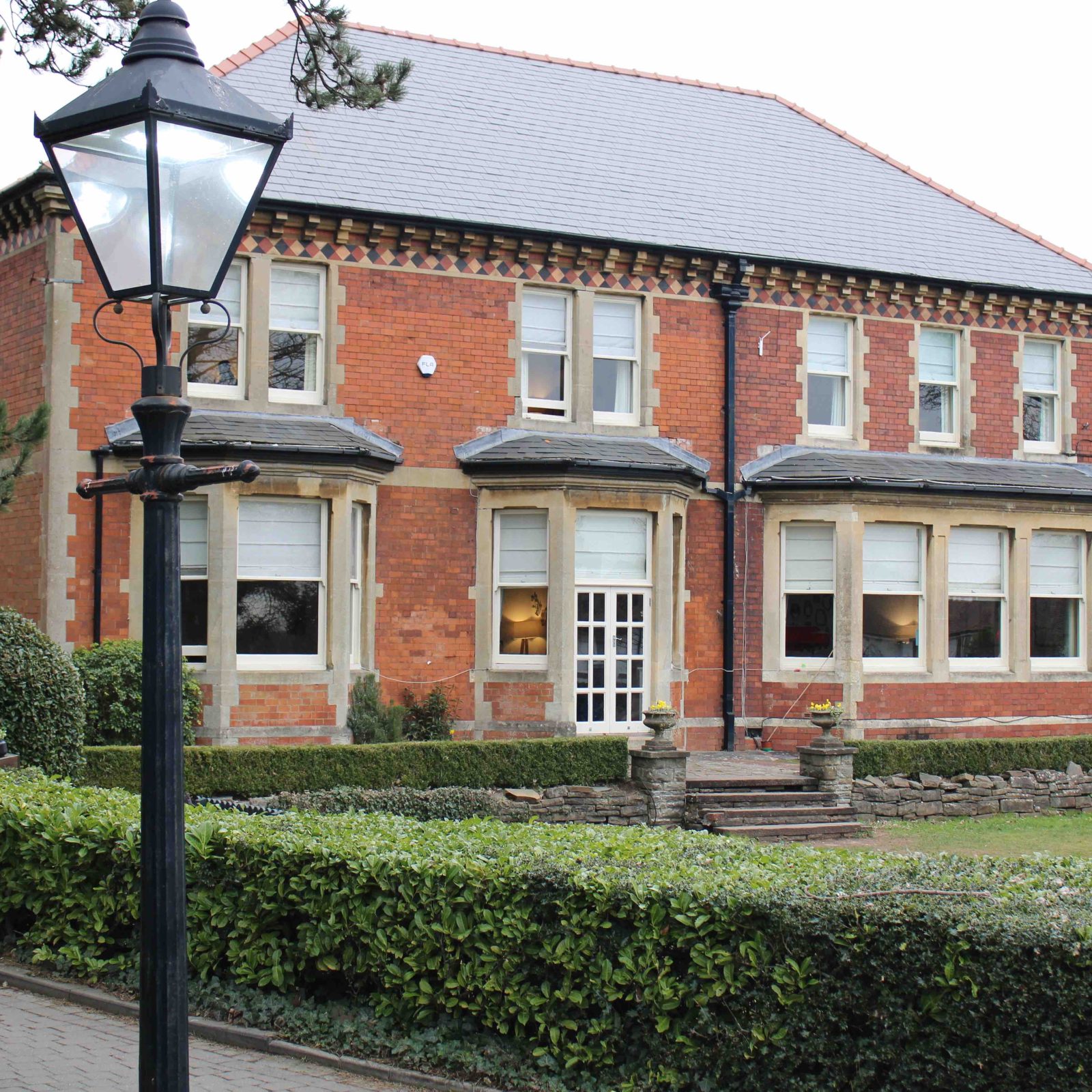 Acorns Nurseries A charming two-story brick house with multiple windows, nestled in the lush greenery of Llanishen, surrounded by hedges and trees. A streetlamp stands gracefully in the foreground amidst fallen acorns, enhancing the picturesque scene.