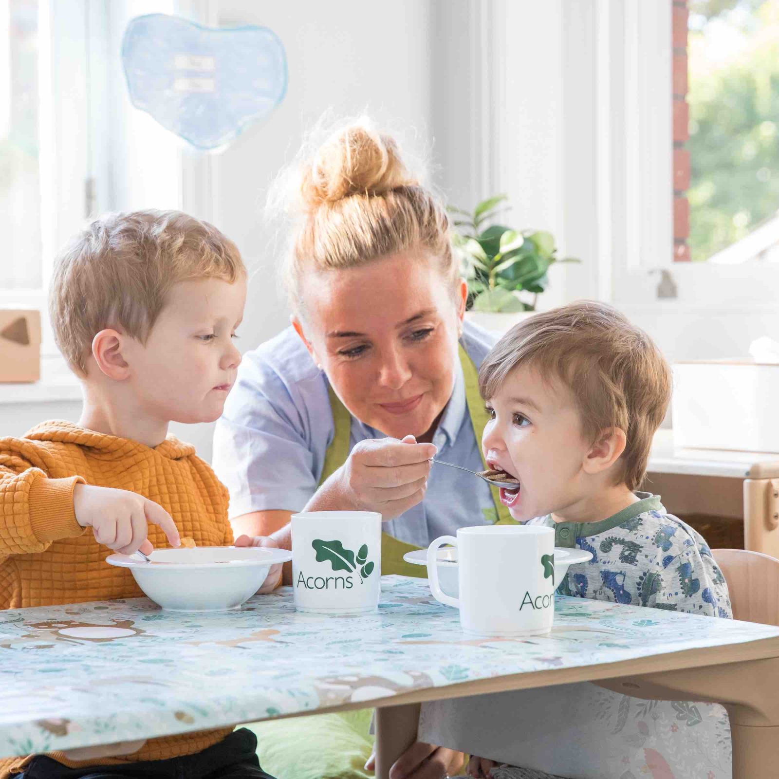 Acorns Nurseries In the warmth of their home, a caregiver feeds two young boys breakfast at a table, with cups labeled 