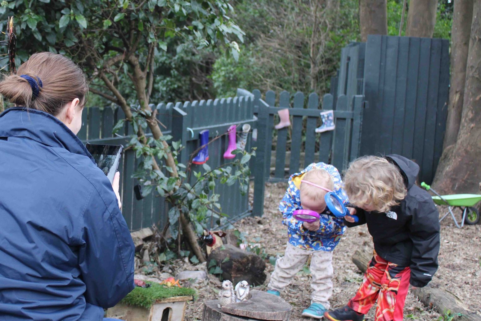 Acorns Nurseries In the comfort of their home yard, an adult captures a photo of two children eagerly examining the ground with magnifying glasses, surrounded by trees and a sturdy fence.