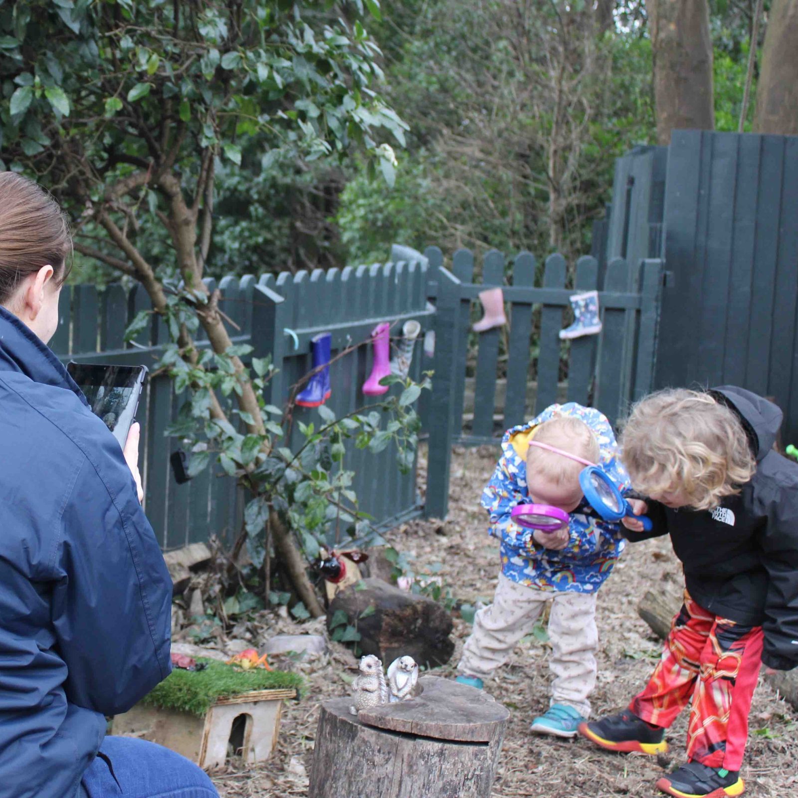 Acorns Nurseries In the comfort of their home yard, an adult captures a photo of two children eagerly examining the ground with magnifying glasses, surrounded by trees and a sturdy fence.