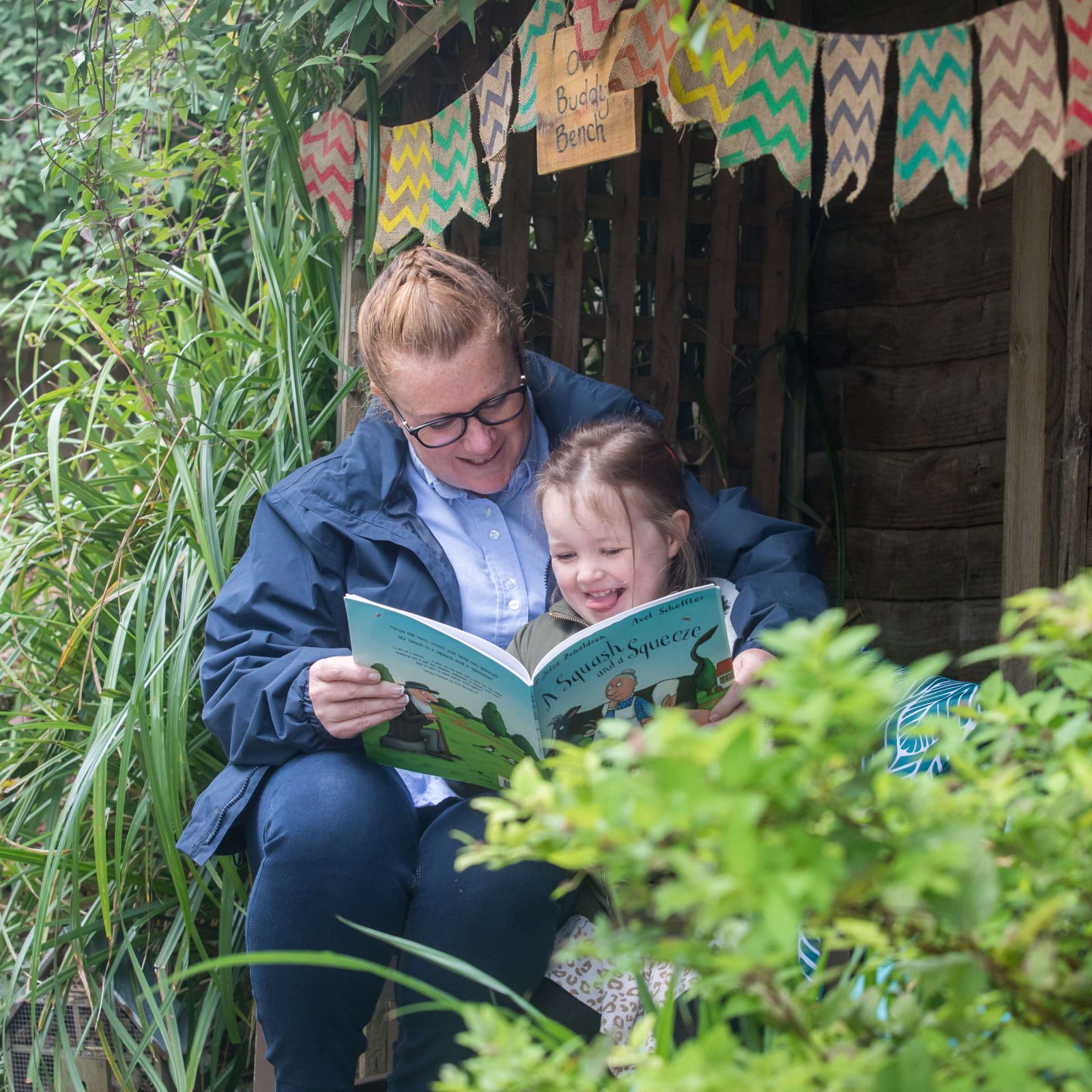 Acorns Nurseries An adult and child sit outdoors reading a book together, surrounded by the cozy charm of home, with lush greenery and colorful bunting enhancing the serene atmosphere.