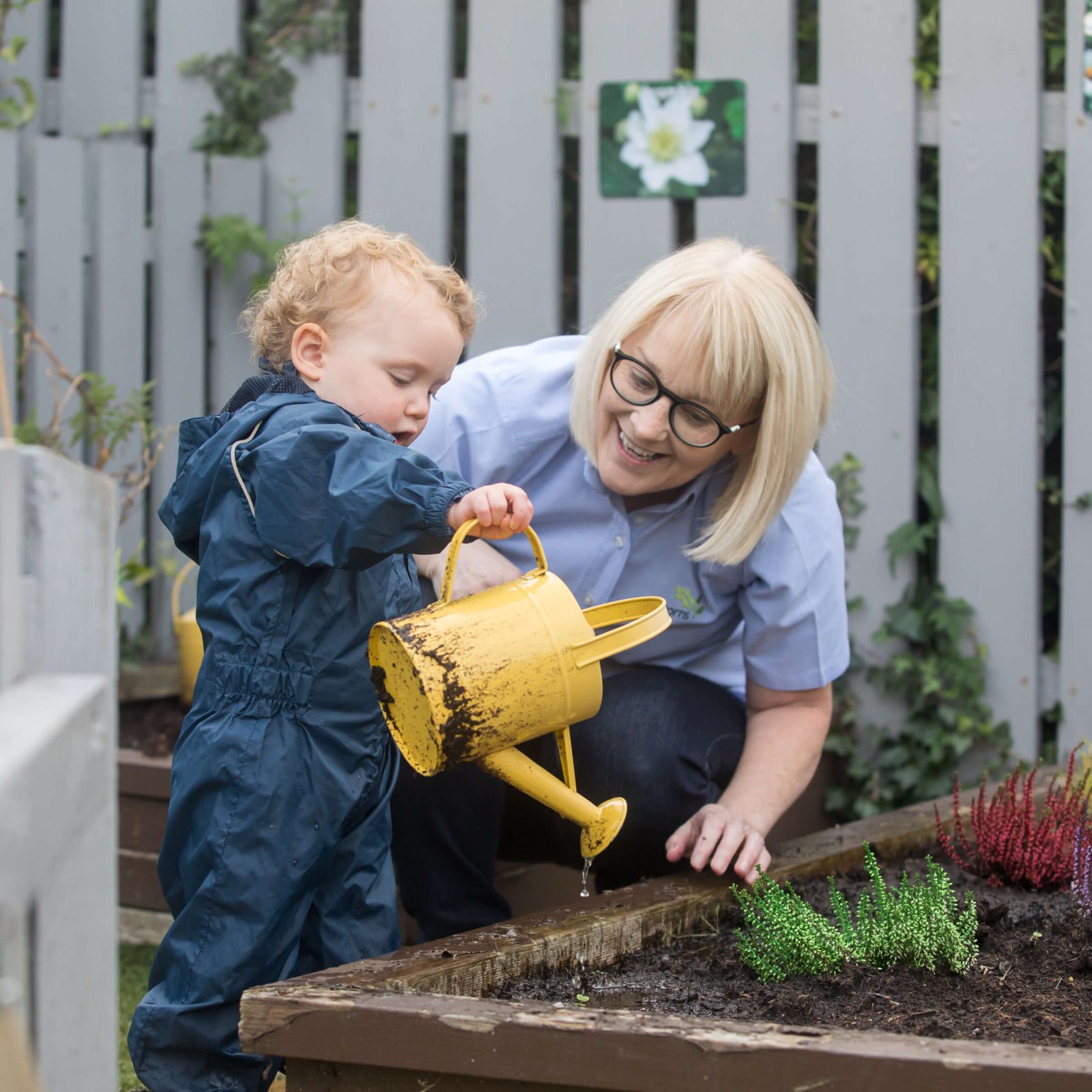 Acorns Nurseries A child waters plants in a garden bed with a yellow watering can, assisted by an adult kneeling beside them in the cozy setting of their home.