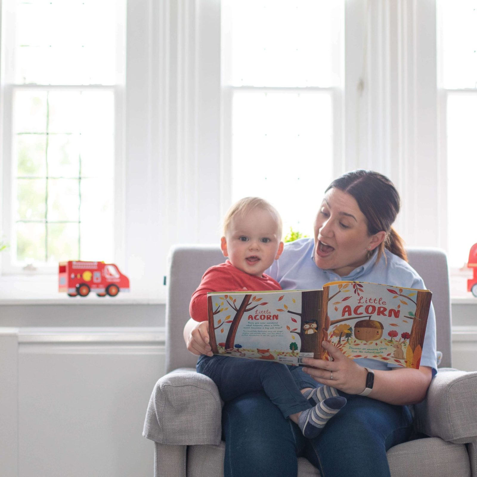 Acorns Nurseries In the comfort of their home, an adult and a child sit in a chair, immersed in a book together. Sunlight spills into the bright room, illuminating toy buses lined up neatly on the windowsill.