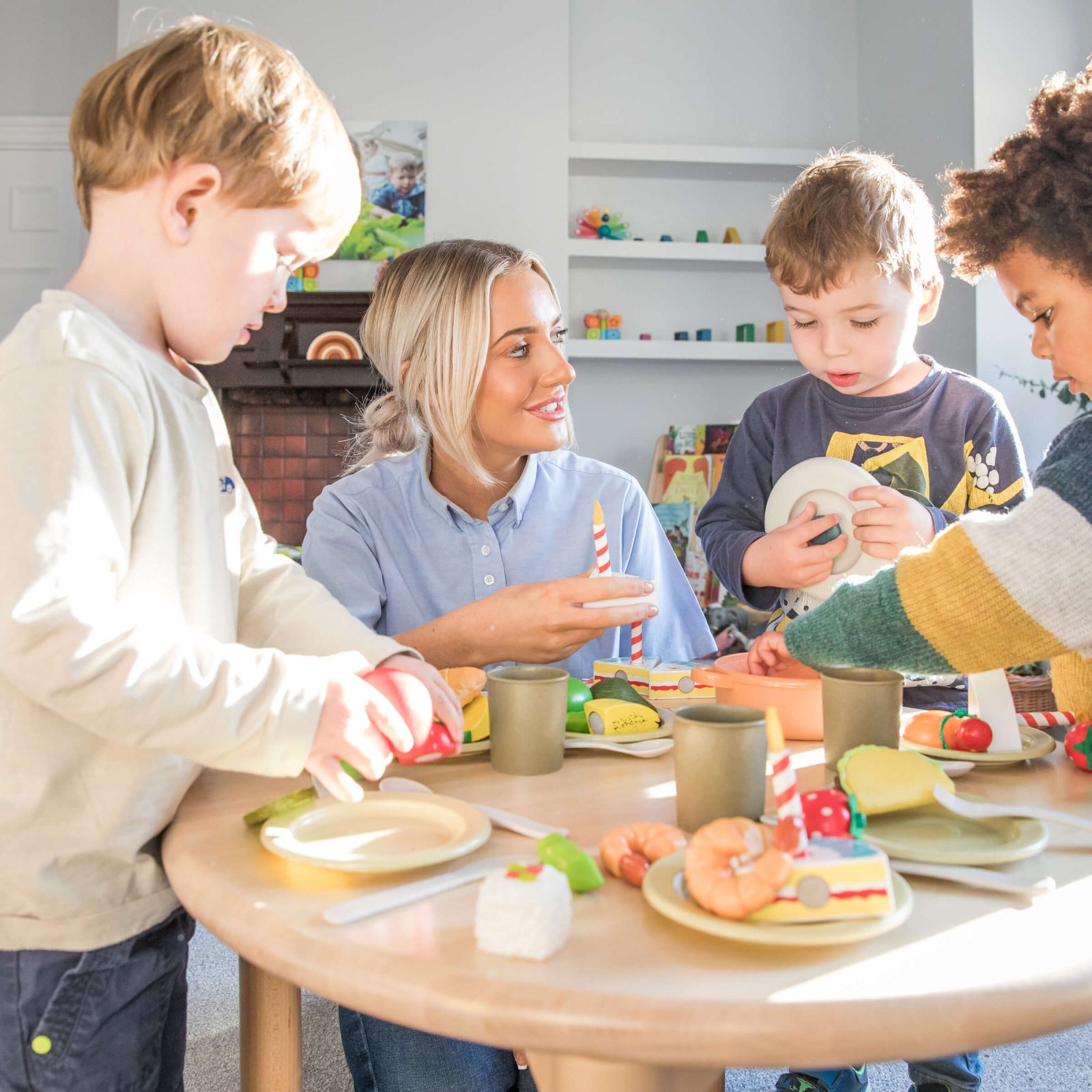 Acorns Nurseries Four children play with toy food at a table, enjoying a warm atmosphere that feels just like home, under the watchful eye of an adult.