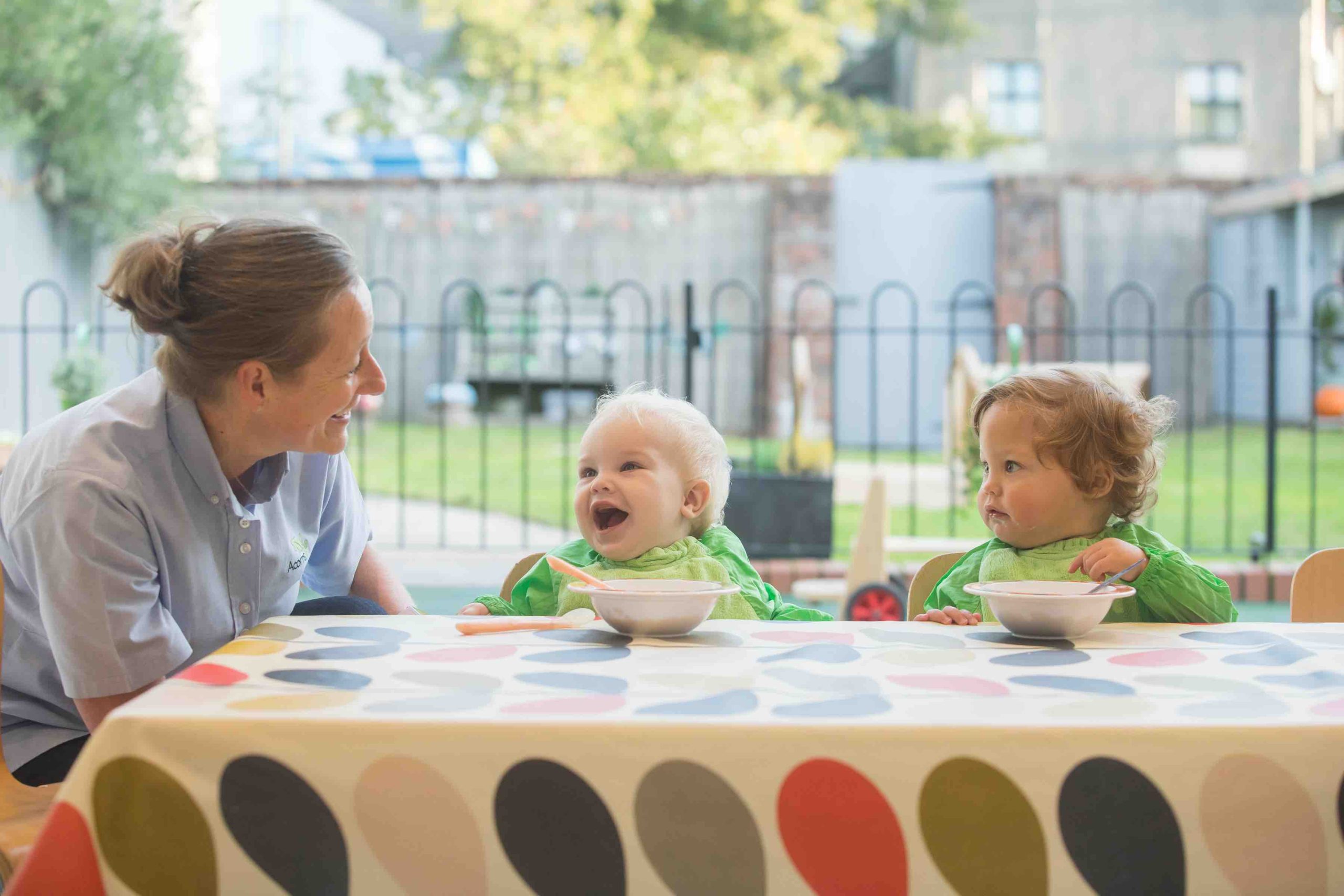 Acorns Nurseries Two toddlers in green bibs sit at a table with a smiling adult. One toddler is laughing, while the other looks at their bowl of healthy food, enjoying the eating experience.